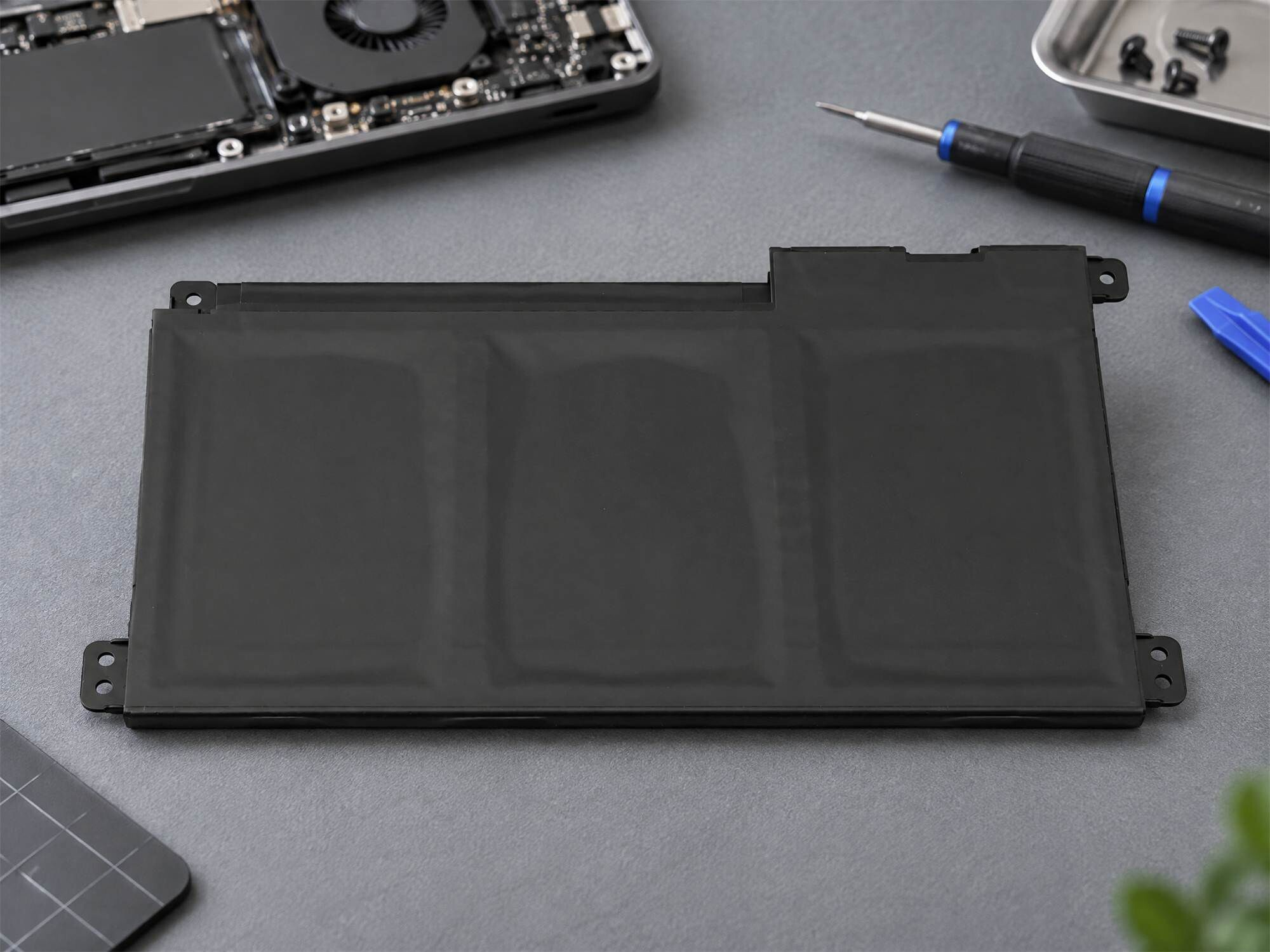 Eine schwarze Laptop-Batterie liegt auf einem grauen Tisch mit Werkzeugen und Laptop-Teilen.