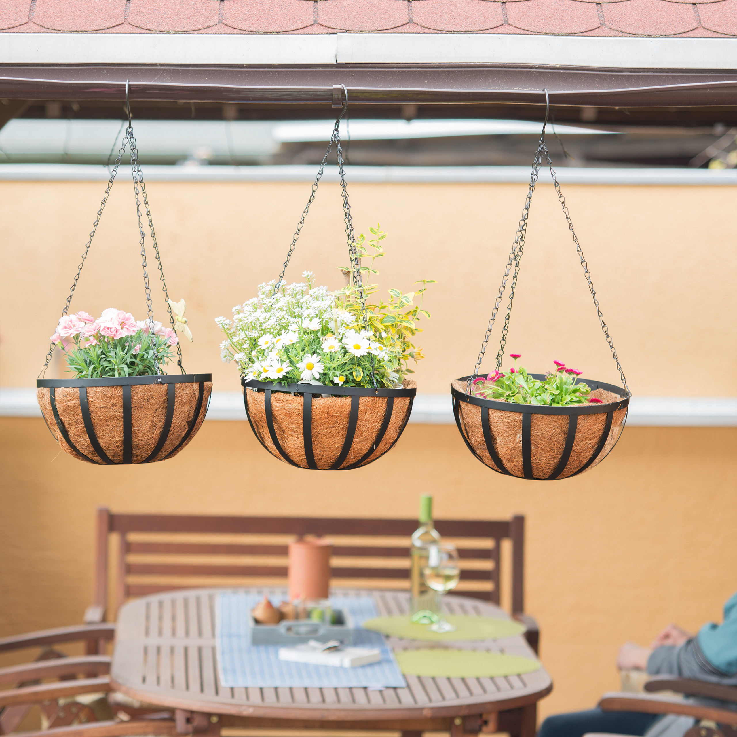 Drei hängende Blumenkörbe mit rosa und weißen Blüten unter einem Vordach, im Hintergrund ein Holztisch.
