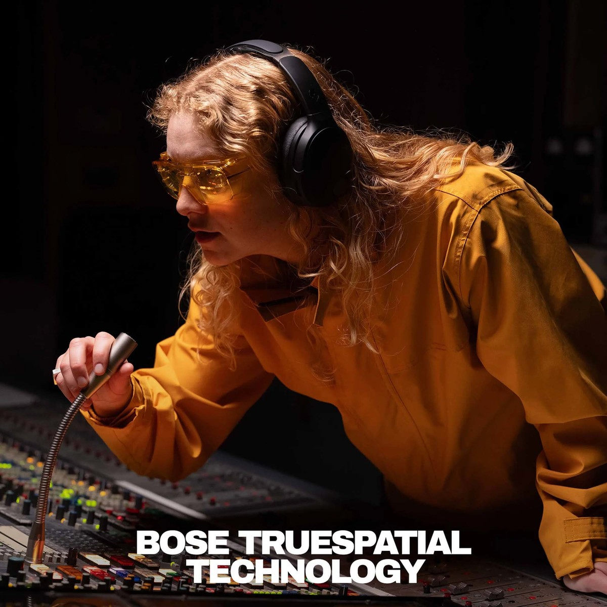 Una persona con gafas amarillas y auriculares ajusta una mesa de mezclas. Texto: BOSE TRUESPATIAL TECHNOLOGY.