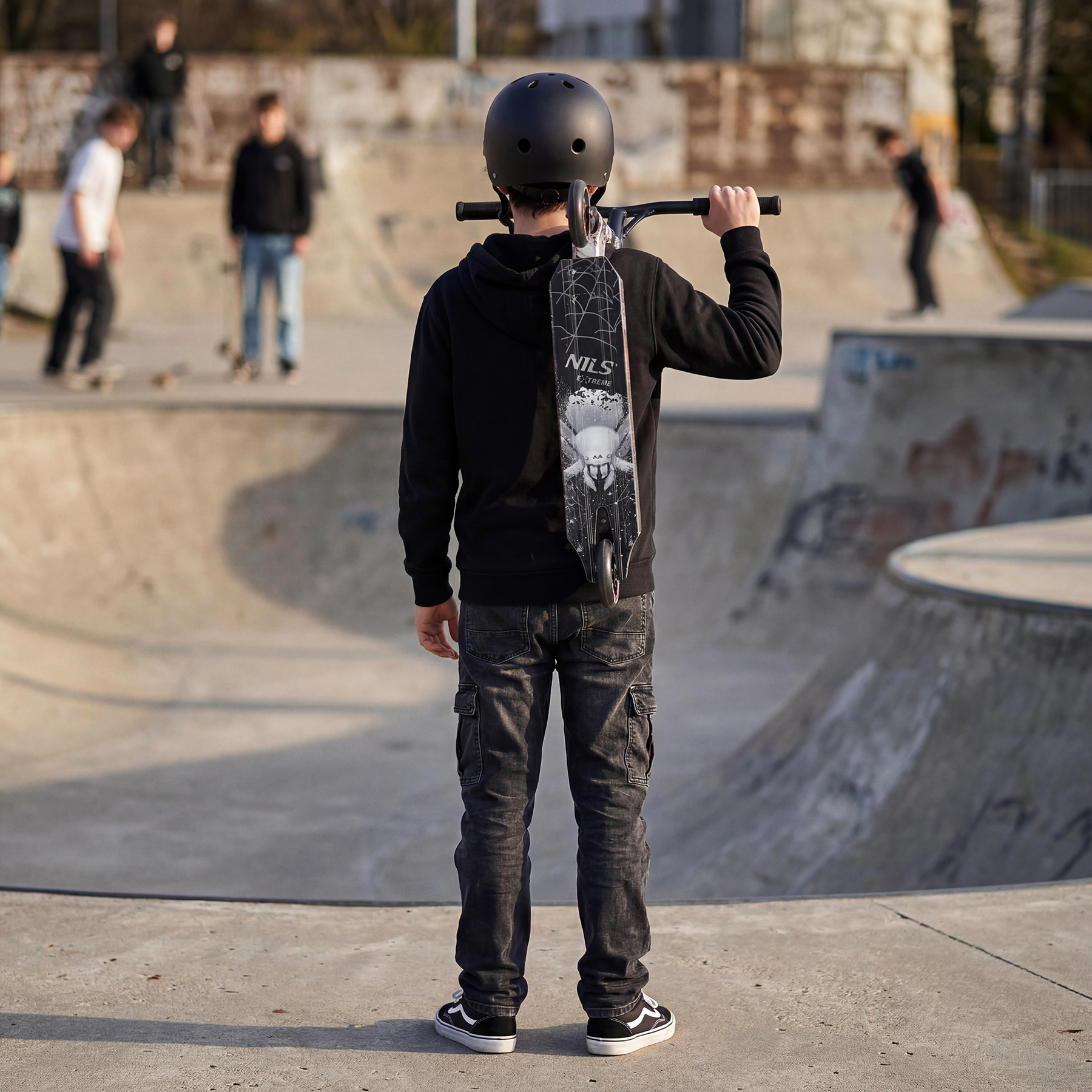 Chłopiec w kasku i czarnej bluzie stoi w skateparku, trzymając hulajnogę wyczynową na ramieniu.