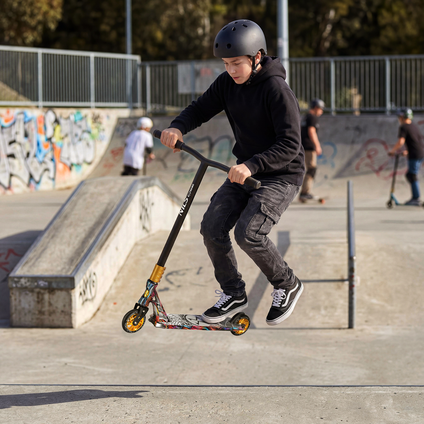 Chłopak w czarnej bluzce z kapturem i kasku wykonuje skok na hulajnodzie wyczynowej w skateparku.