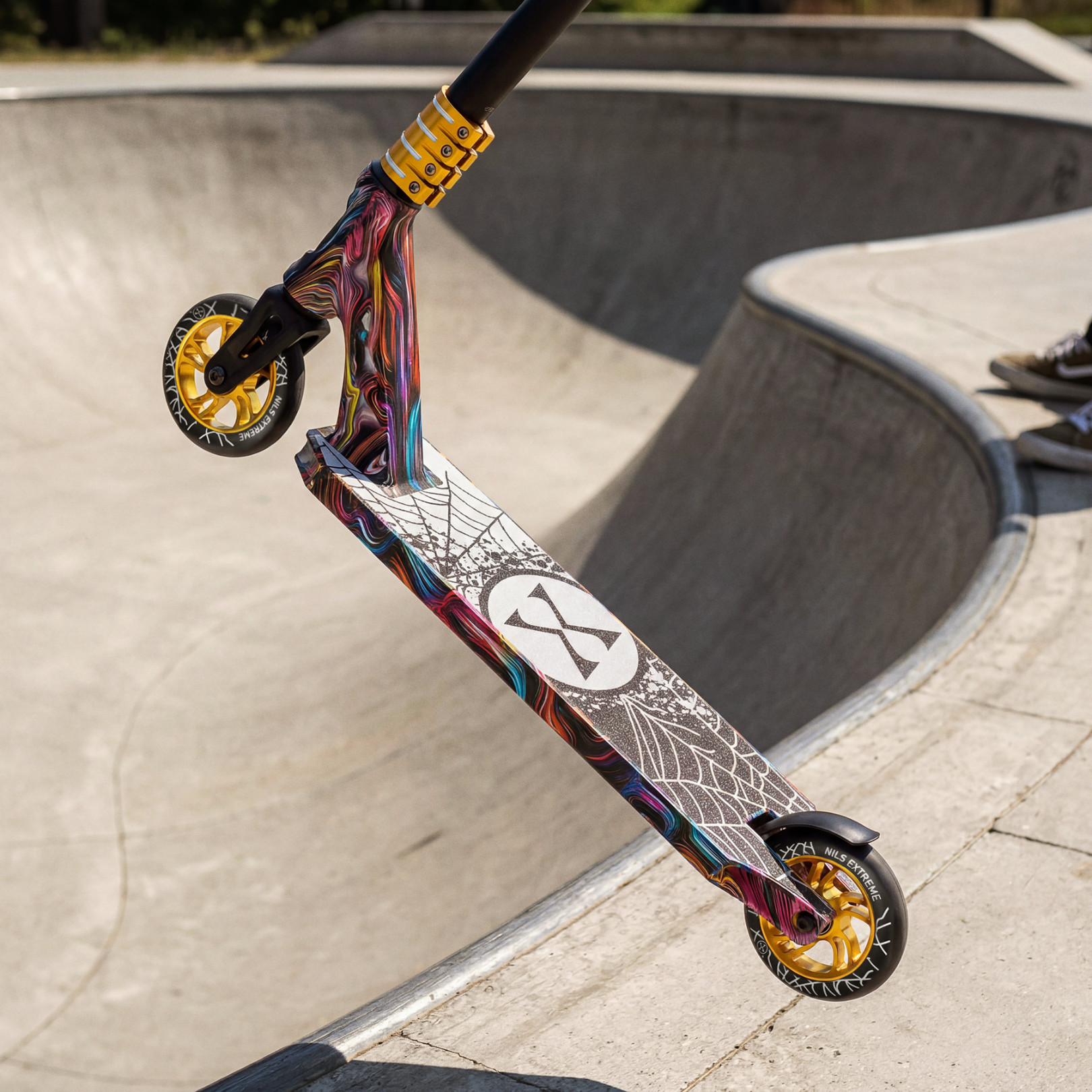Kolorowa hulajnoga wyczynowa ze złotymi felgami i grafiką pajęczyny unosi się nad rampą skateparku.
