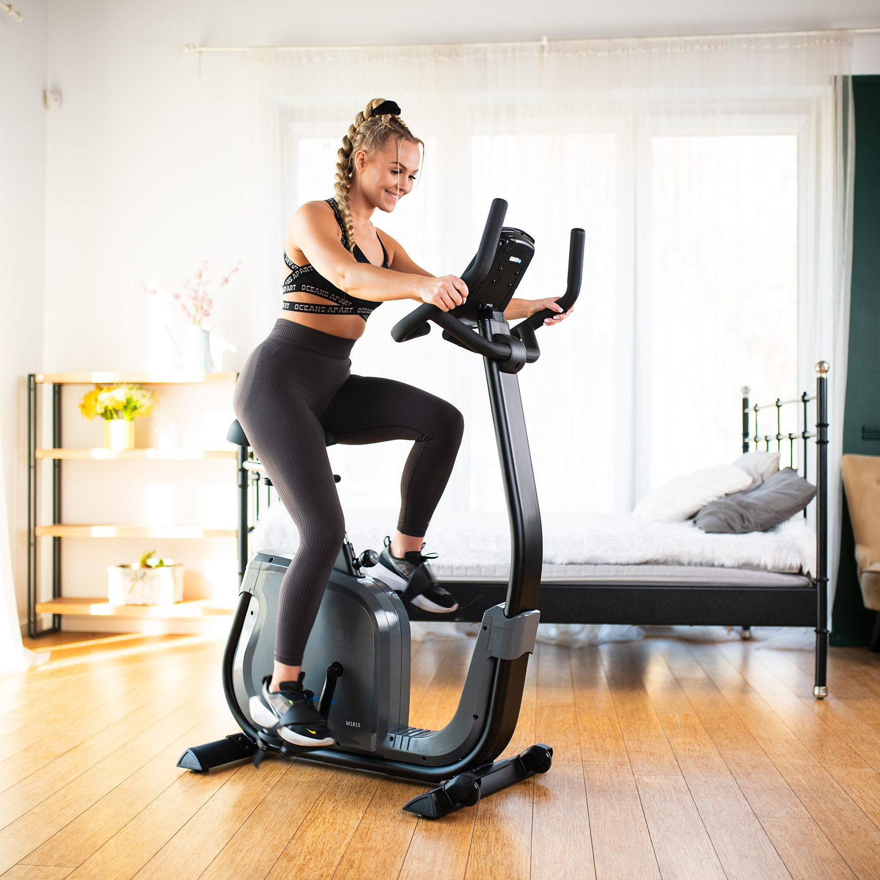 Blond kobieta w stroju sportowym ćwiczy na rowerku stacjonarnym w jasnej sypialni z drewnianą podłogą.
