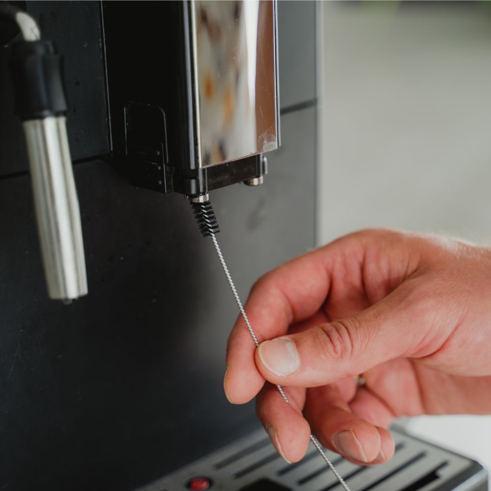 A hand cleans the coffee machine spout with a small cleaning brush.