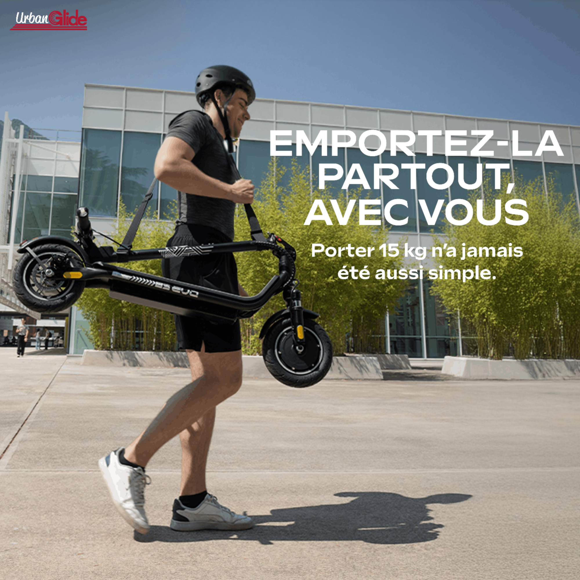 Un hombre con casco lleva un patinete eléctrico plegado con una correa frente a un edificio moderno.