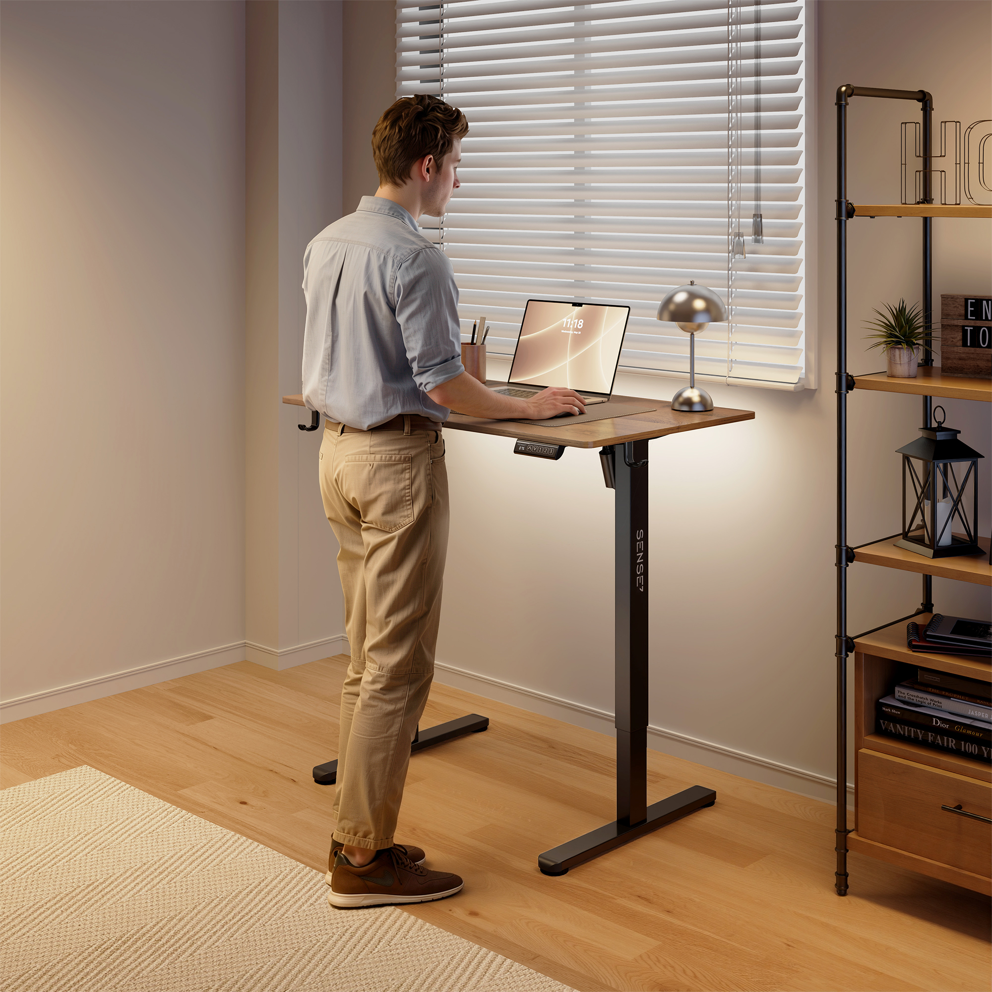 Un hombre de pie en un escritorio ajustable de madera usa una laptop en una habitación luminosa con persianas.
