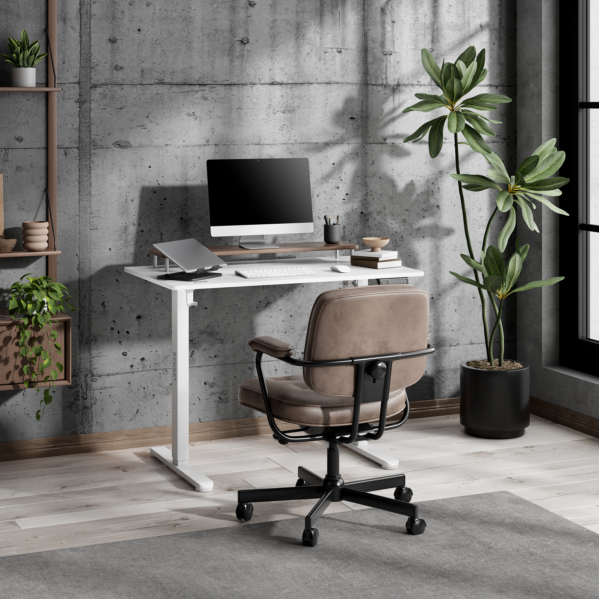 Oficina moderna con escritorio blanco, silla de cuero marrón, ordenador y grandes plantas verdes ante una pared de hormigón.