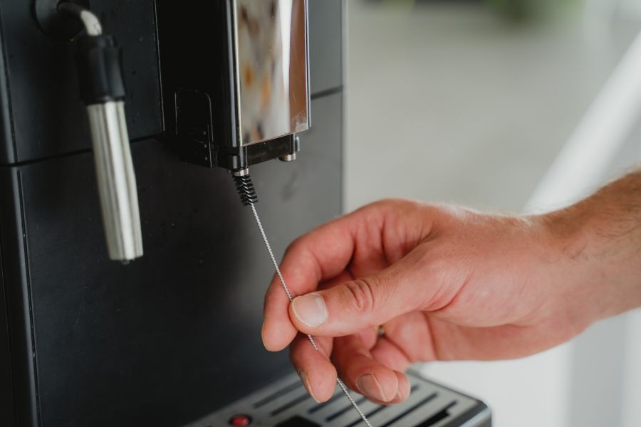 A hand cleaning the coffee machine nozzle with a small brush.