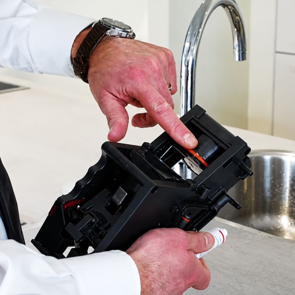A person in a white shirt points at a part inside a black coffee machine component near a kitchen sink.