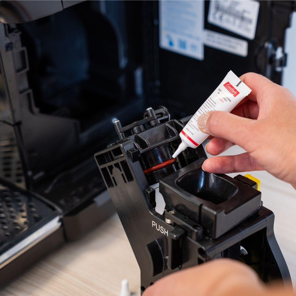 A hand applies lubricant from a small tube onto the red O-ring of a coffee machine brew group.