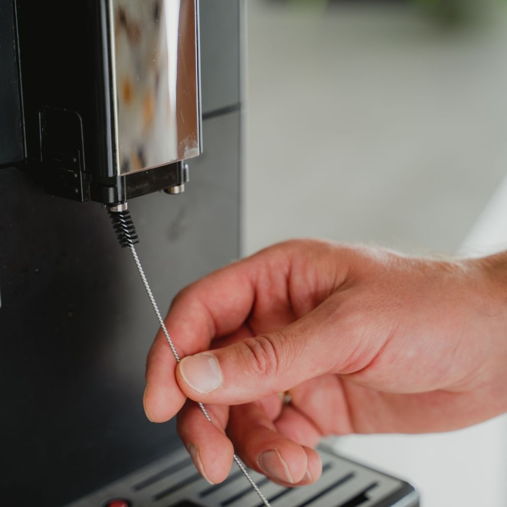 A hand cleans a coffee machine's nozzle with a small wire brush.