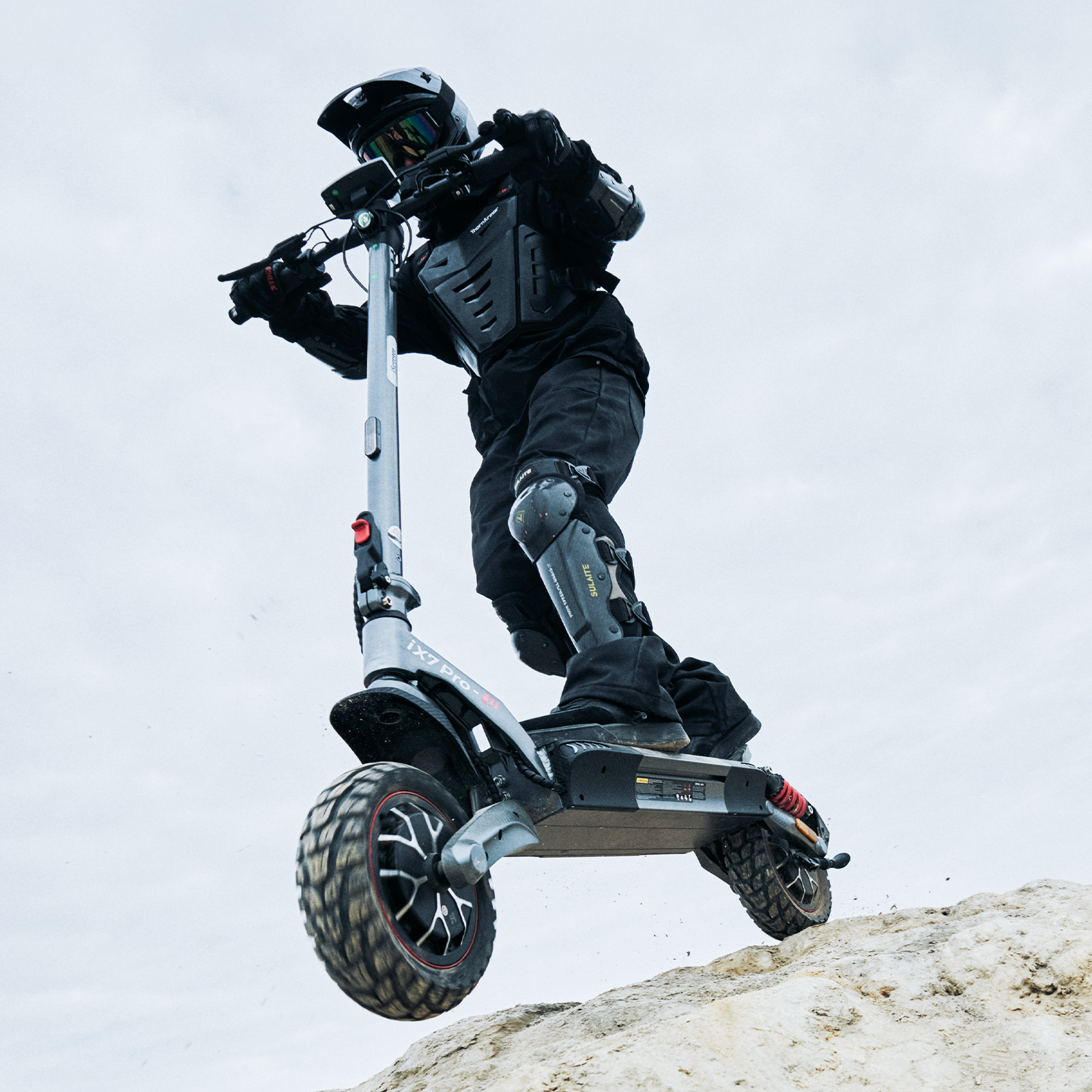 Un piloto de motocross con casco y equipo de protección salta en un patinete eléctrico sobre una cresta de arena.