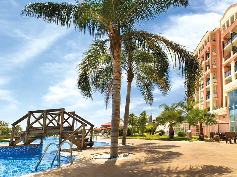 Una zona de piscina soleada con un pequeño puente de madera, palmeras, tumbonas y un gran hotel al fondo.