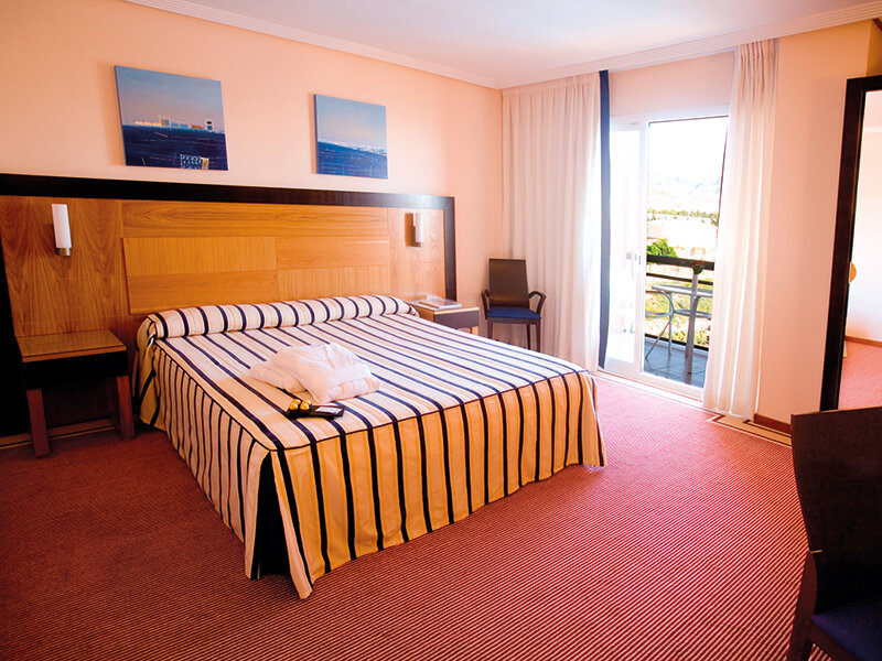 Una habitación de hotel luminosa con una cama grande, manta a rayas, cabecero de madera y puerta al balcón.