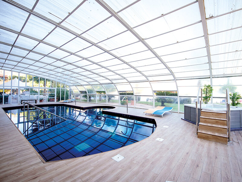 Piscina cubierta bajo techo abovedado de vidrio, con jacuzzi, terraza de madera y tumbonas.