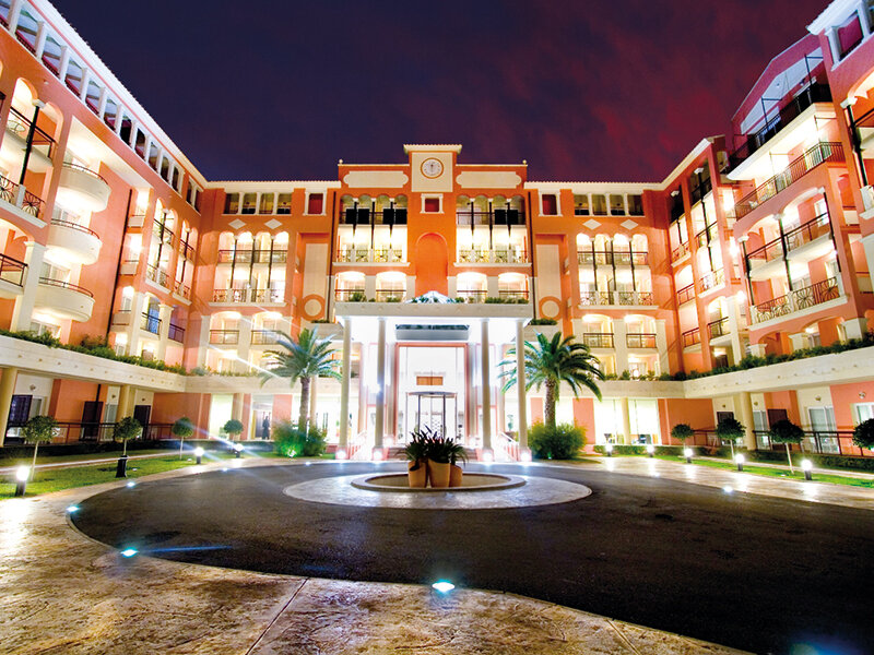 Una gran fachada de hotel naranja iluminada por la noche con una entrada circular y palmeras.