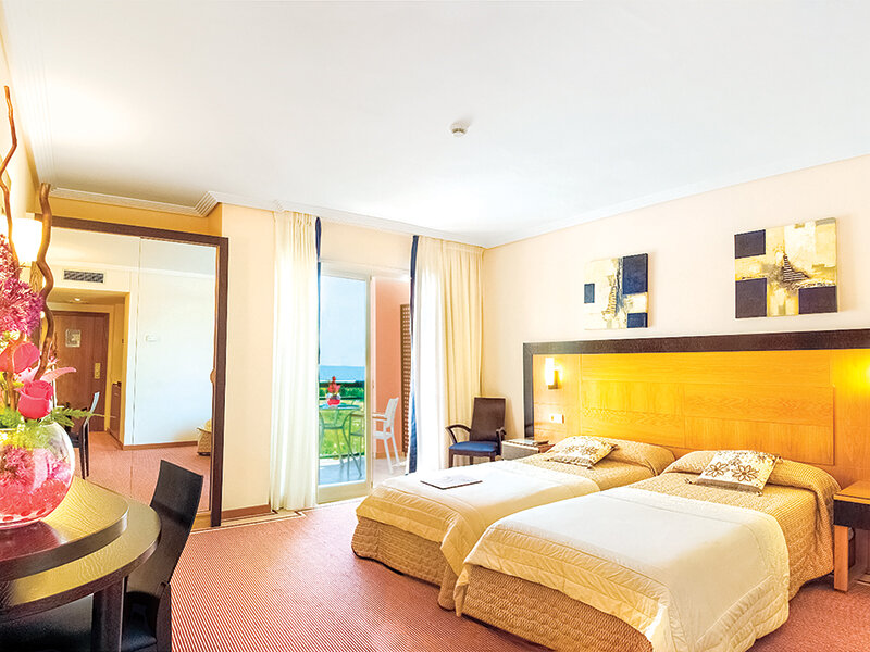 Una habitación de hotel luminosa con dos camas, un gran espejo y una puerta de balcón con vista al exterior.