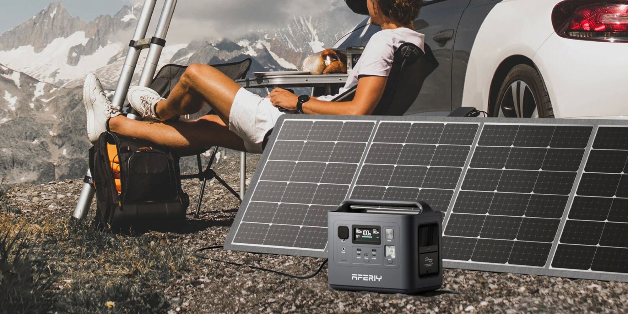 Un panel solar portátil y una estación de energía en primer plano, con una persona sentada junto a un auto ante montañas nevadas.
