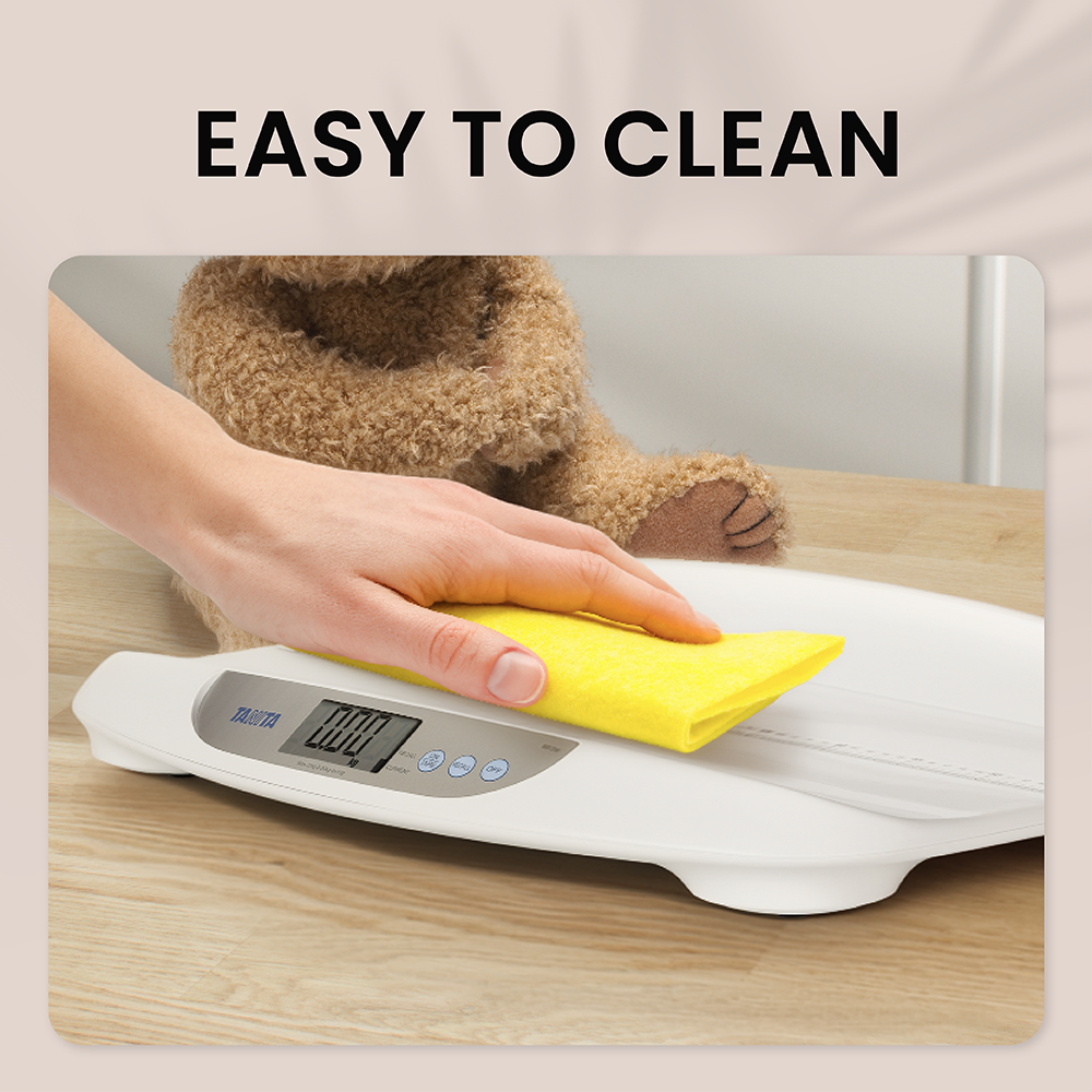 A hand wipes a white baby scale with a yellow cloth, with a teddy bear sitting in the background.