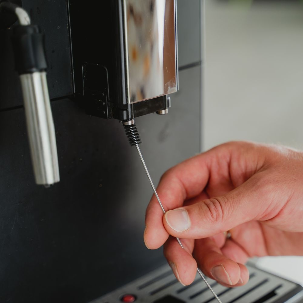 A hand cleans a coffee machine nozzle with a small brush.