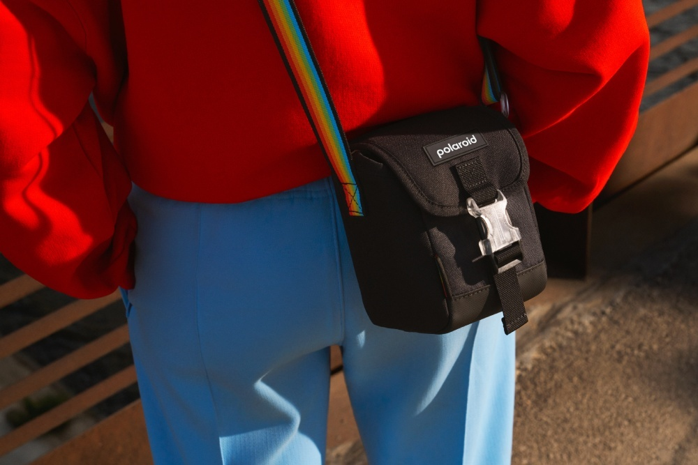 Person in rotem Pullover und blauen Hosen trägt eine schwarze Polaroid-Tasche mit Regenbogenriemen.