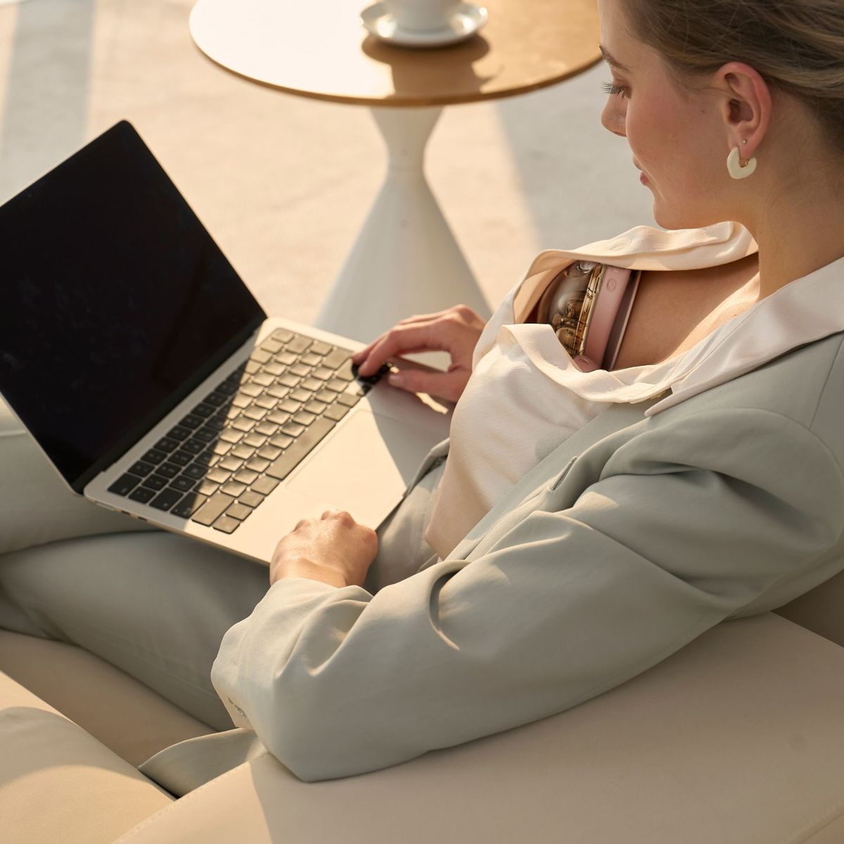 Eine Frau im hellen Blazer sitzt auf einem Sofa und benutzt einen Laptop auf ihrem Schoß.