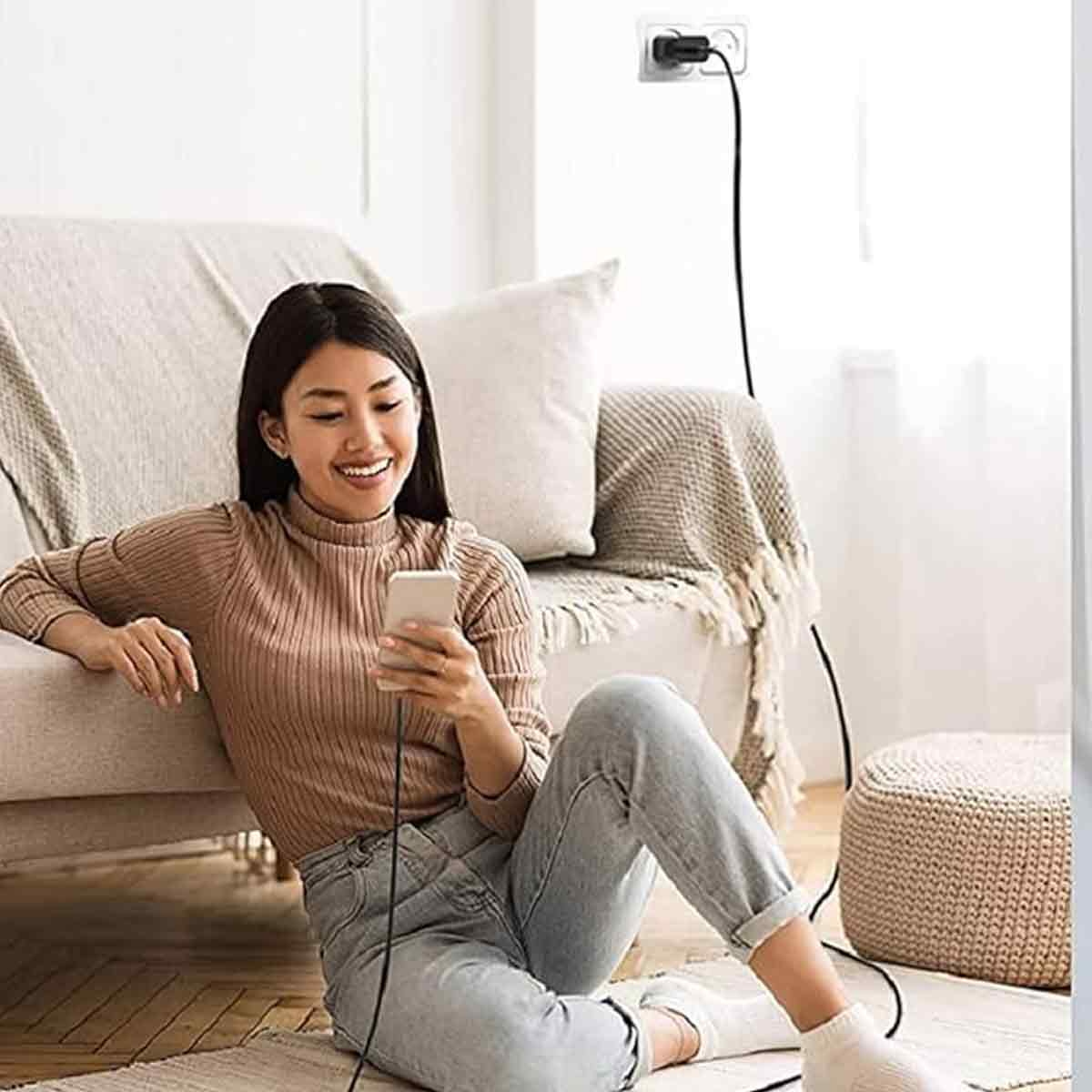 Eine Frau sitzt auf einem Teppich vor einem Sofa und hält ein Smartphone, während ein schwarzes Kabel zur Steckdose führt.
