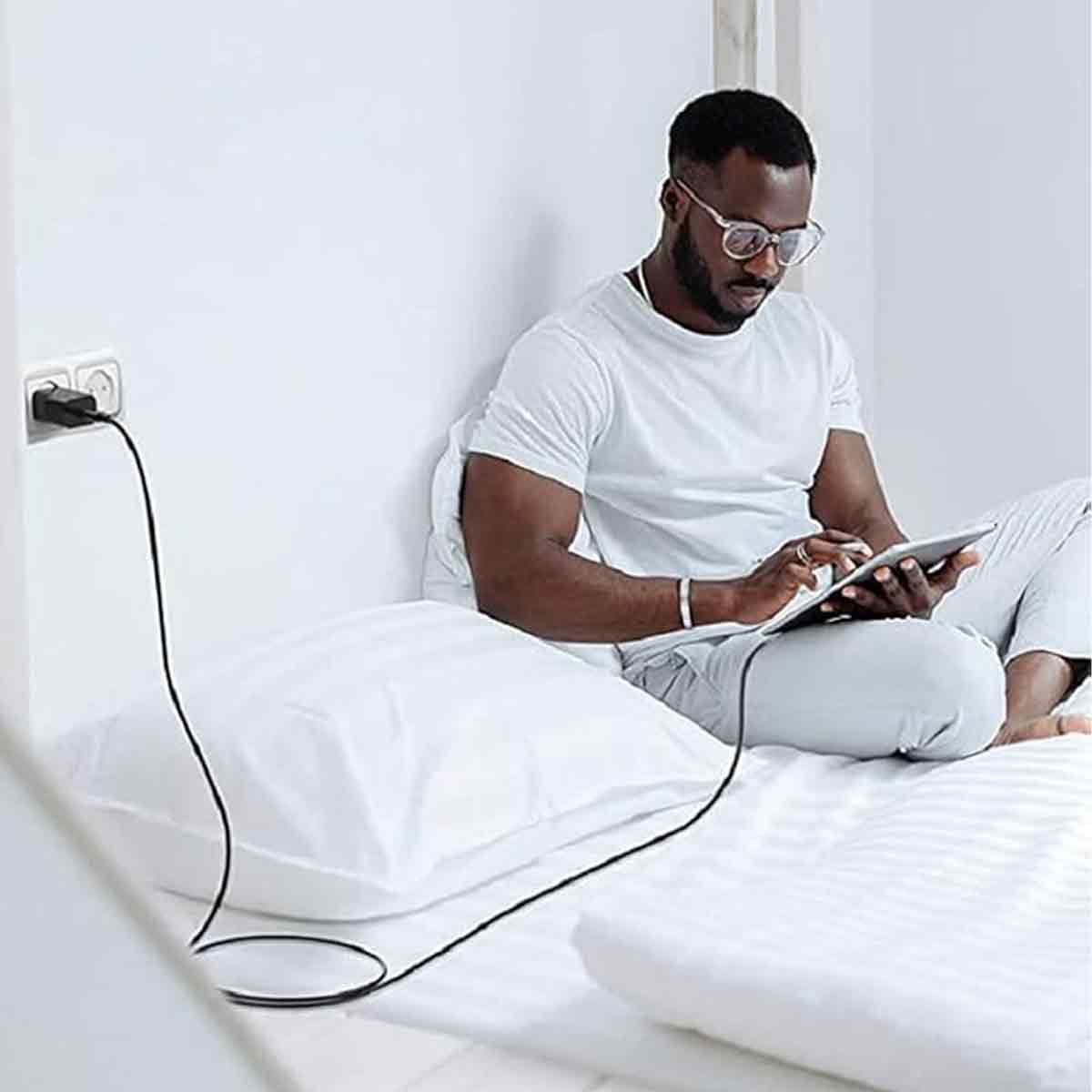 Ein Mann sitzt im Bett und benutzt ein Tablet, während es an einer Steckdose an der Wand angeschlossen ist.
