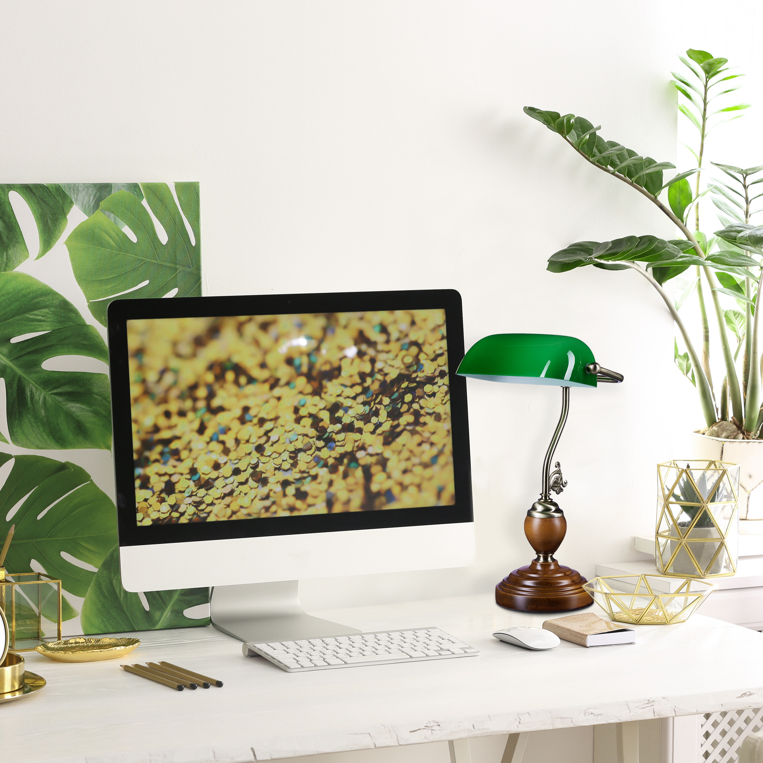 Ein weißer Schreibtisch mit einem Monitor, einer grünen Lampe, goldenen Dekorationen und Topfpflanzen davor.