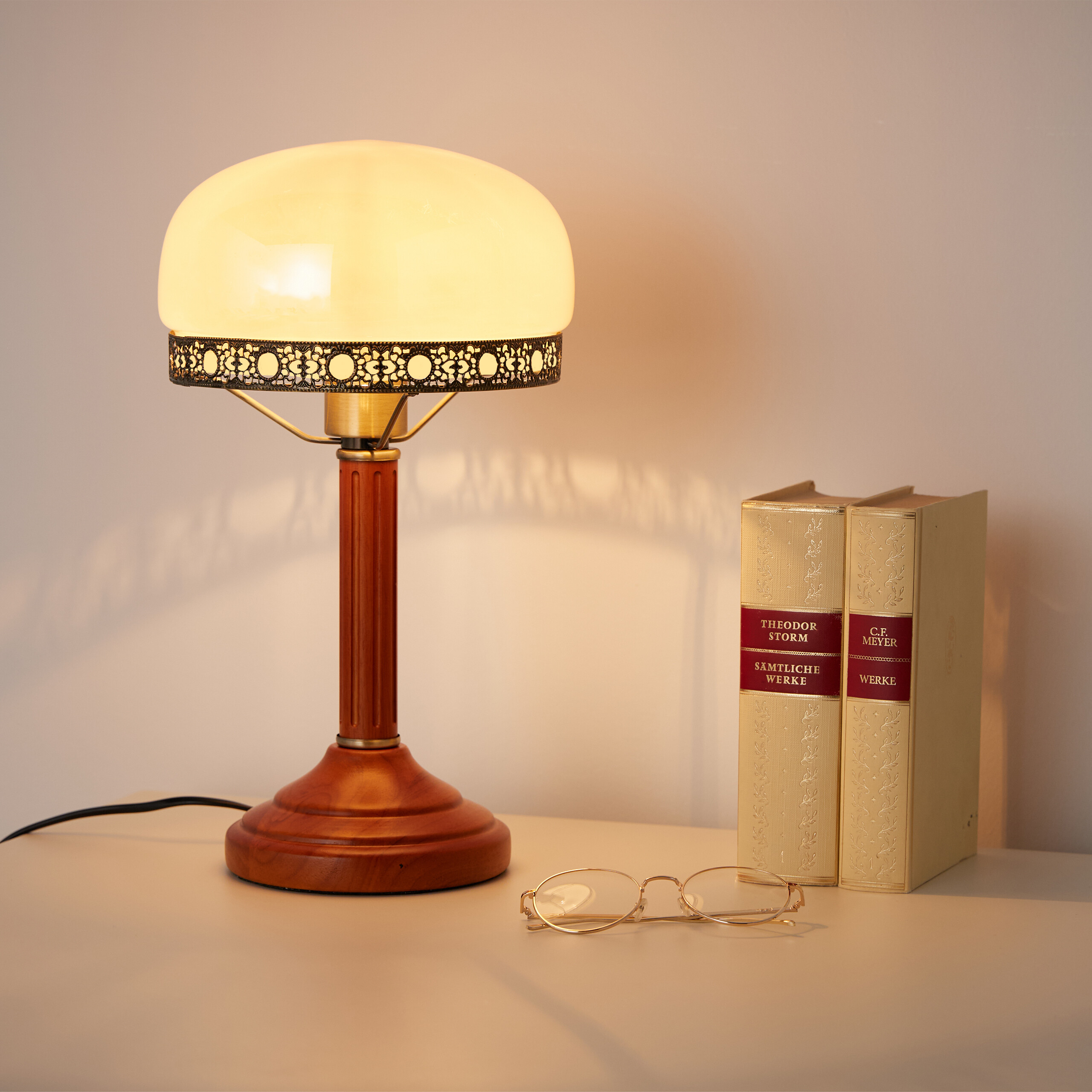 Eine erleuchtete Tischlampe, zwei weiße Bücher und eine Brille stehen auf einem hellen Tisch vor einer grauen Wand.