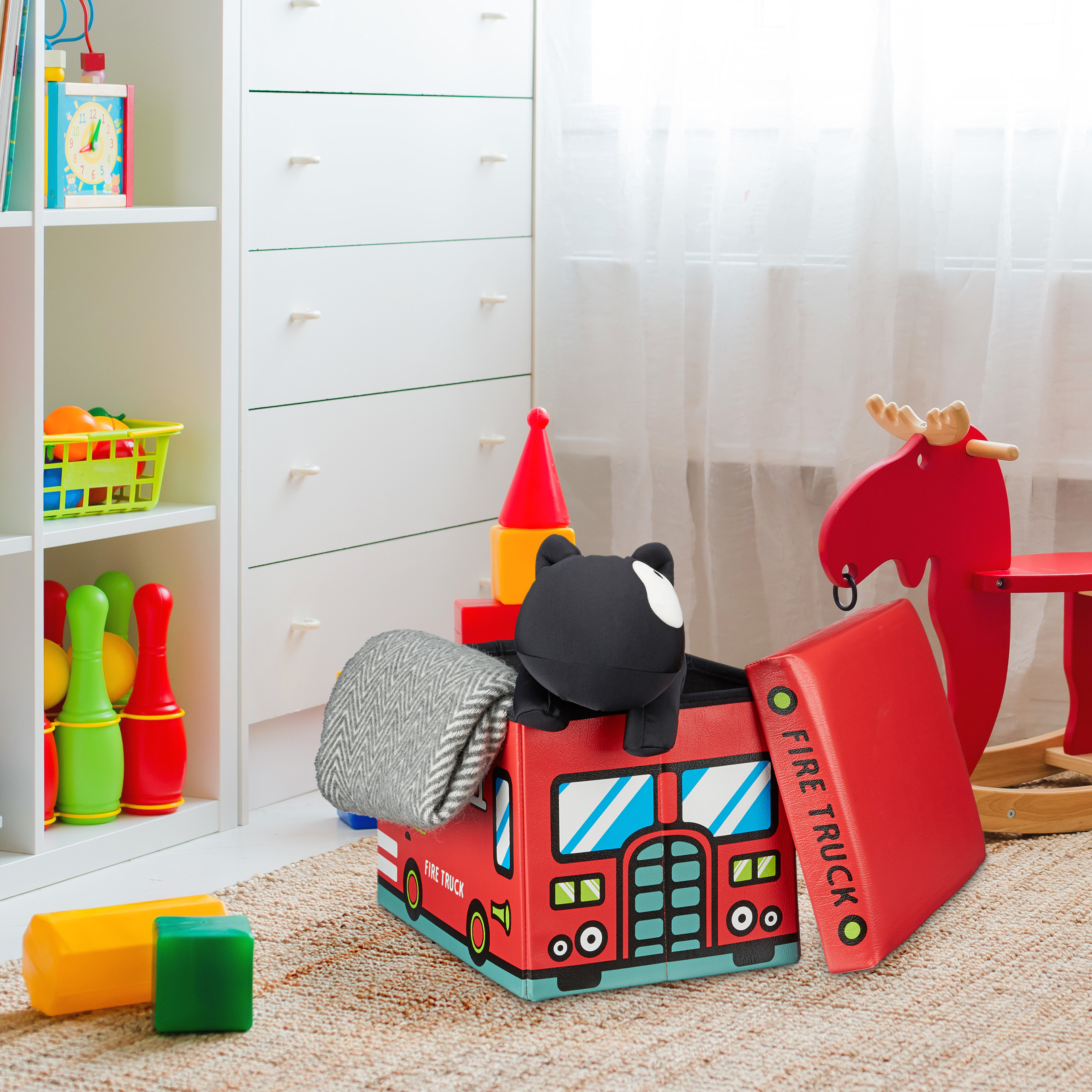 Eine rote Feuerwehr-Spielzeugkiste in einem Kinderzimmer mit Spielzeug, einer weißen Kommode und einem roten Schaukel-Elch.
