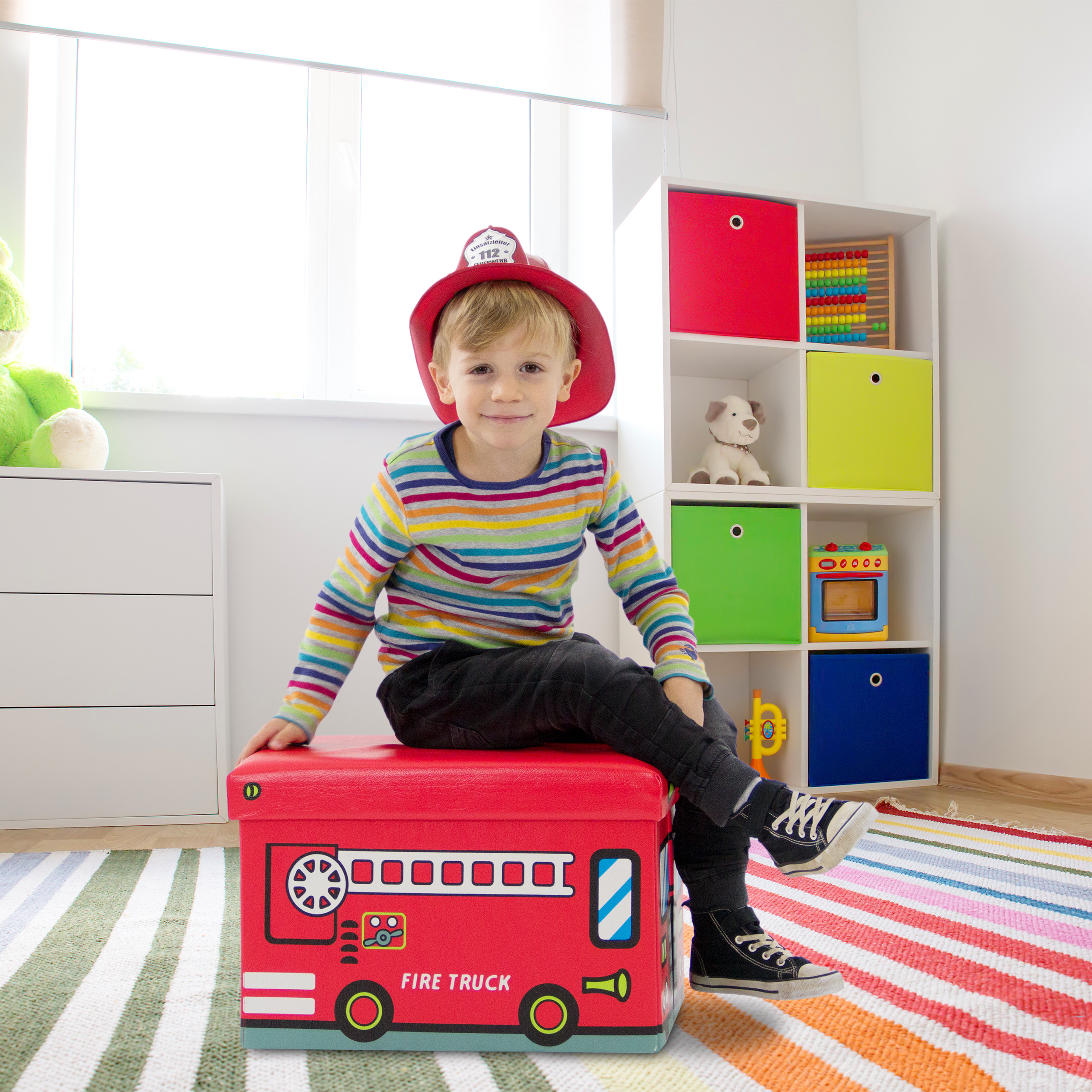 Ein Junge mit Feuerwehrhelm sitzt auf einer roten Spielzeugkiste in einem hellen Zimmer.