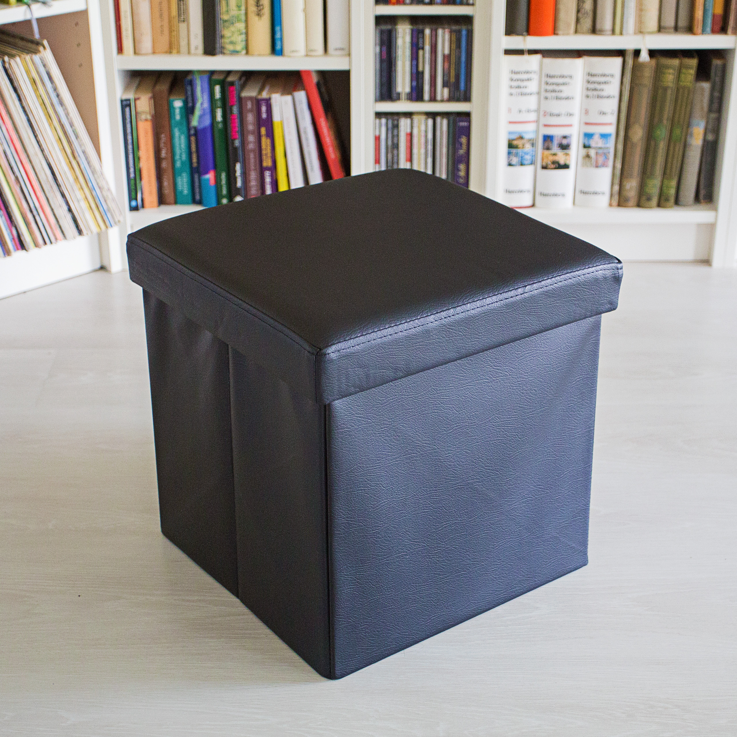Ein schwarzer Leder-Hocker steht auf hellem Boden vor Bücherregalen mit Büchern und CDs.