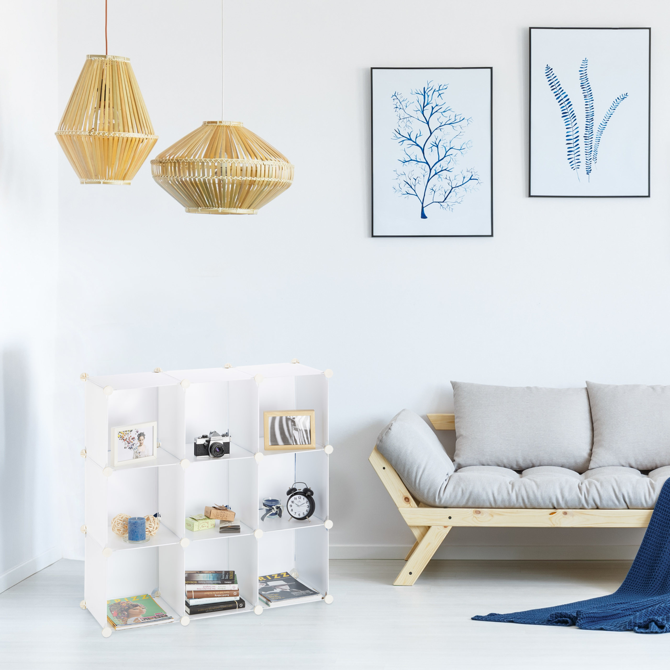 Ein weißes Regal steht neben einem hellen Holzsofa mit grauen Kissen und zwei blauen Pflanzenbildern an der Wand.