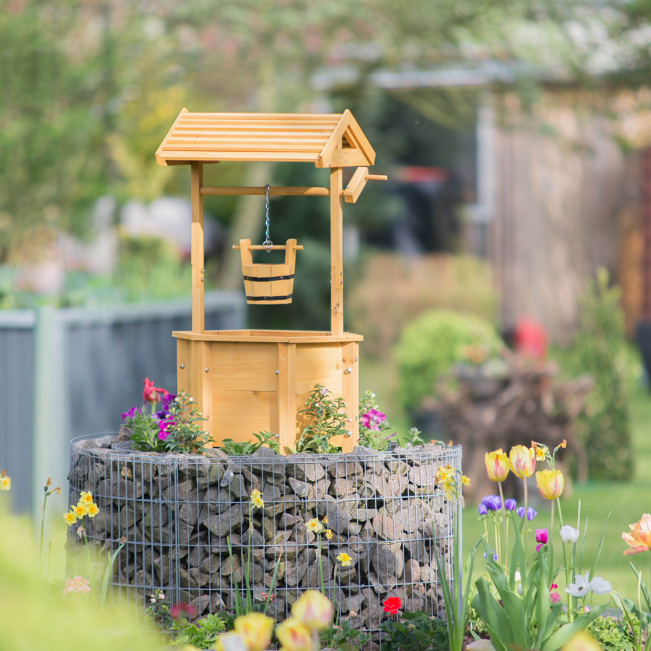 Ein hölzerner Zierbrunnen auf Steinsockel, umgeben von bunten Blumen im Garten.