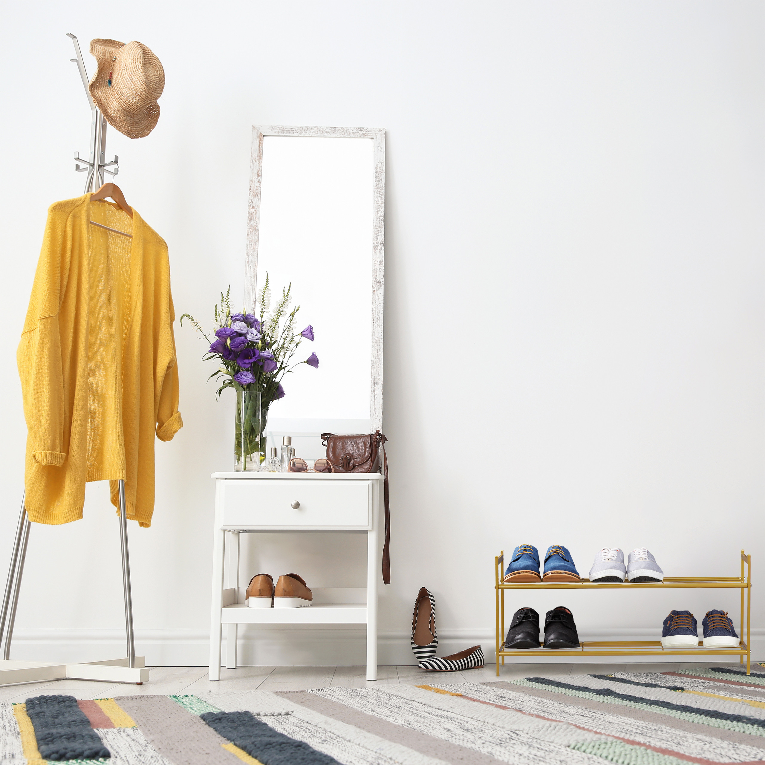 Flur mit Hutablage, gelber Strickjacke, Spiegel, Kommode, Blumen, Schuhen und gestreiftem Teppich vor weißer Wand.