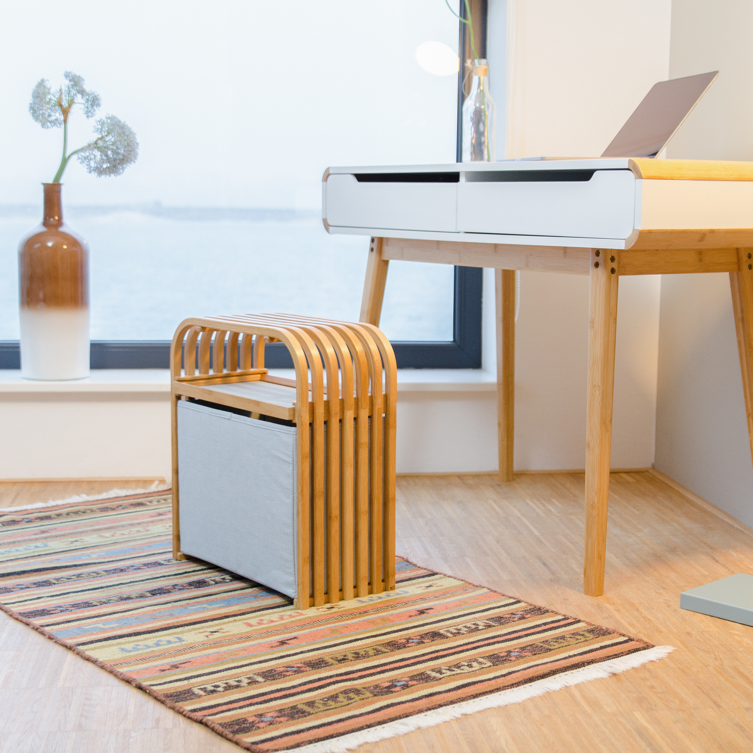 Ein Holztisch, ein Hocker mit grauem Stoff, ein gemusterter Teppich und eine Vase mit Blumen am Fenster.