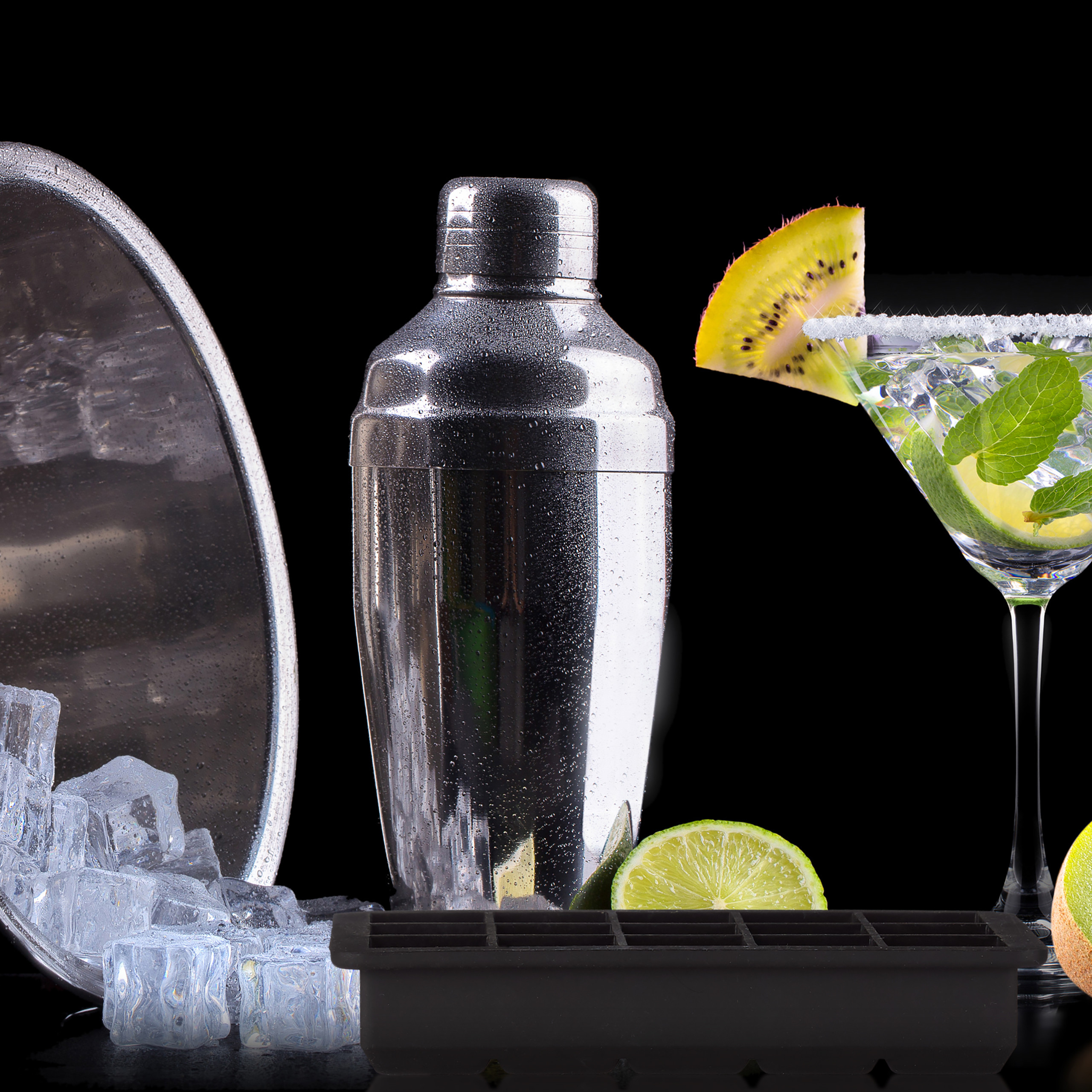 Cocktailshaker, Eiswürfel, Limettenscheiben und ein Martini-Glas mit Kiwi auf schwarzem Hintergrund.