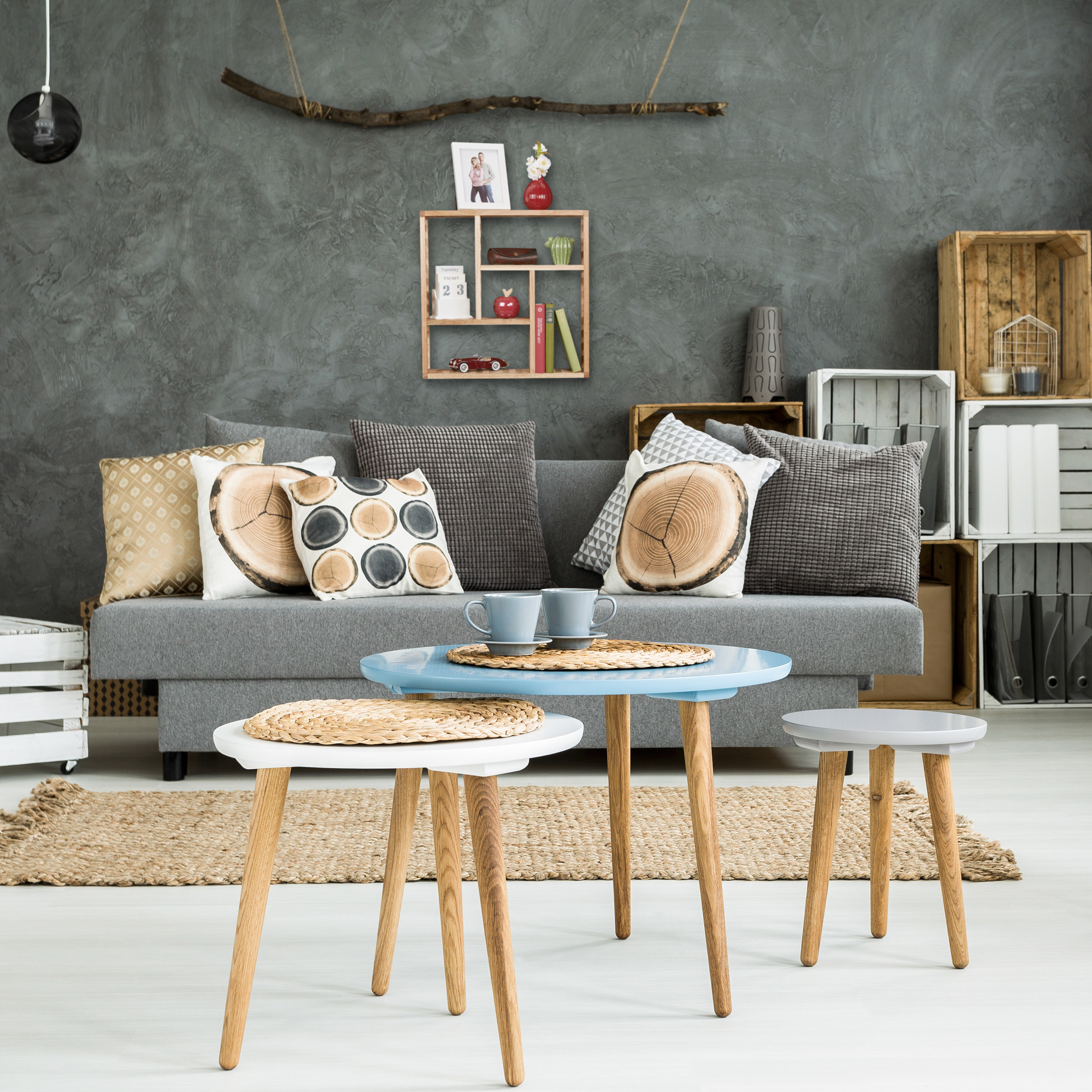 Gemütliches Wohnzimmer mit grauem Sofa, Kissen, blauen und weißen Tischen sowie Holzkisten an der dunklen Wand.