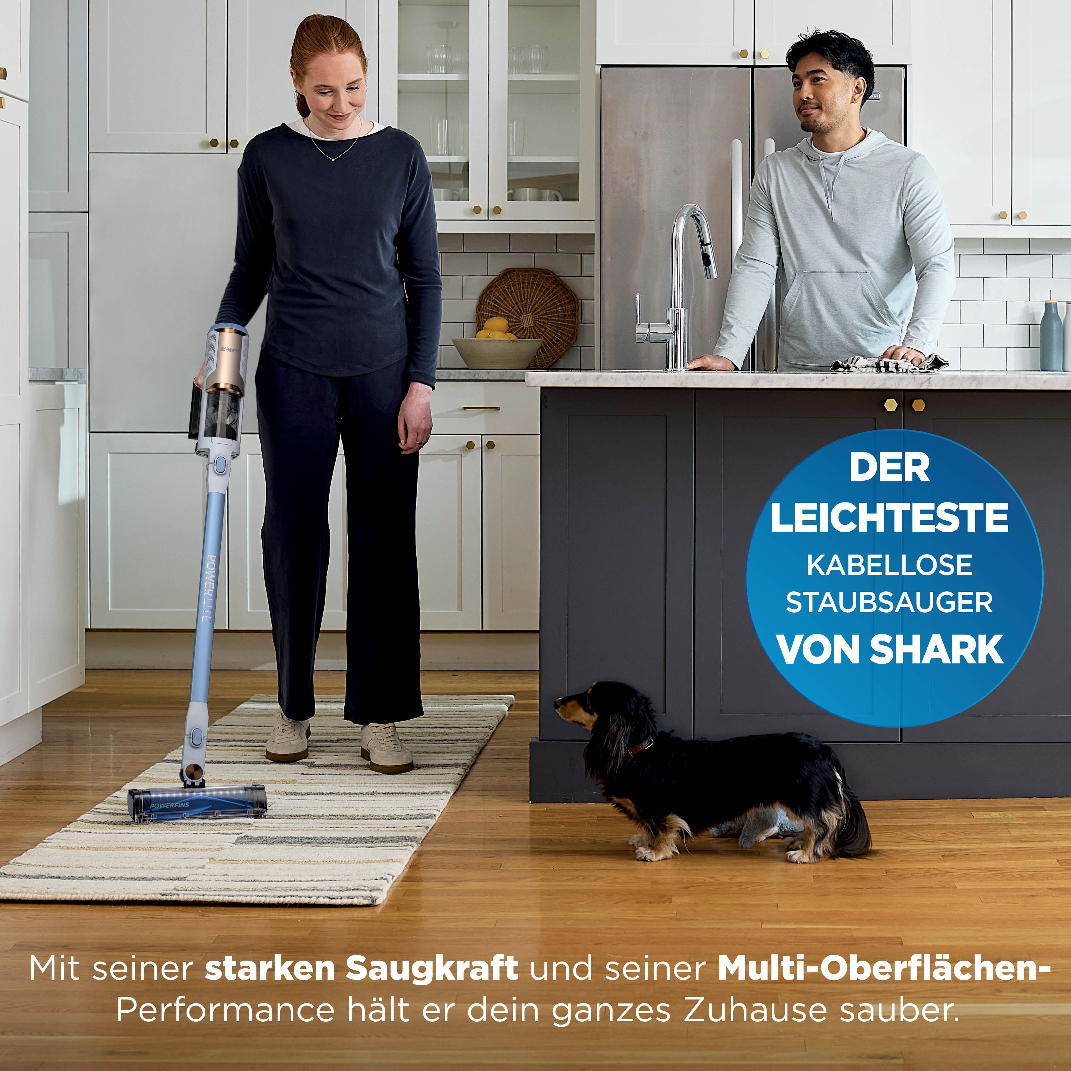 Eine Frau saugt einen Teppich in der Küche, während ein Mann zuschaut und ein Hund daneben steht.