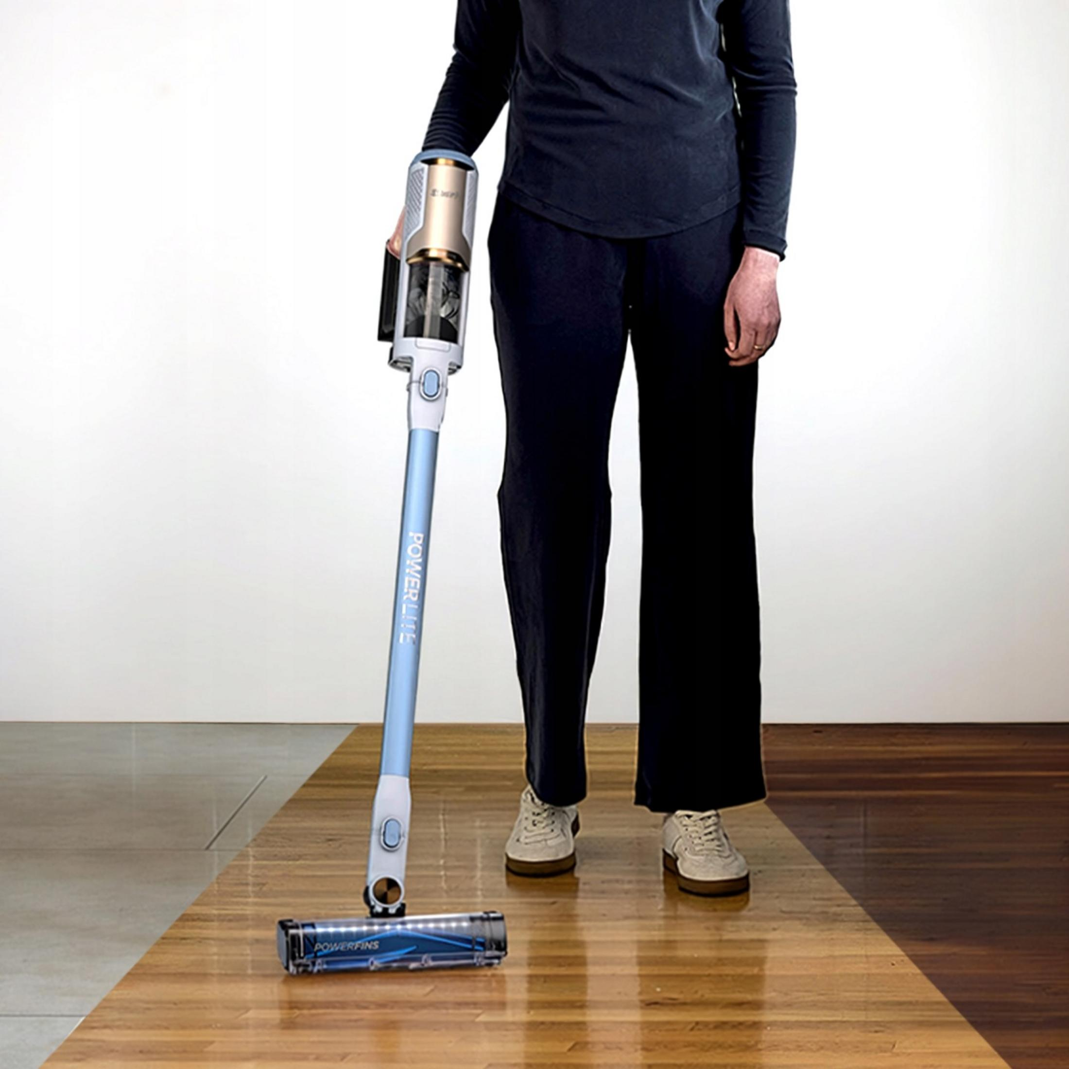 Eine Person im schwarzen Outfit hält einen blauen und silbernen Akkusauger auf einem Holzfußboden.