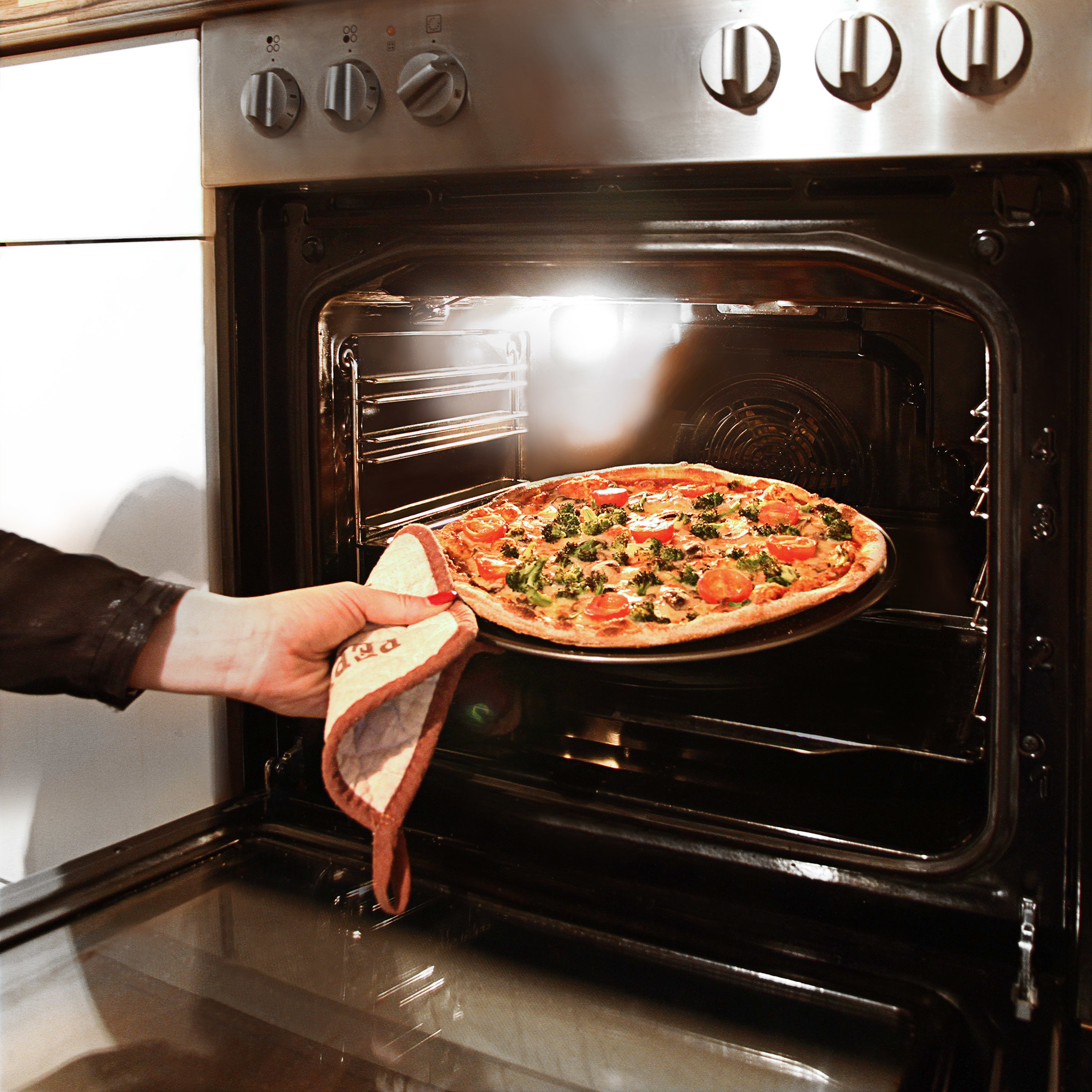 Eine Hand mit Ofenhandschuh nimmt eine Pizza mit Gemüse aus dem heißen Ofen.