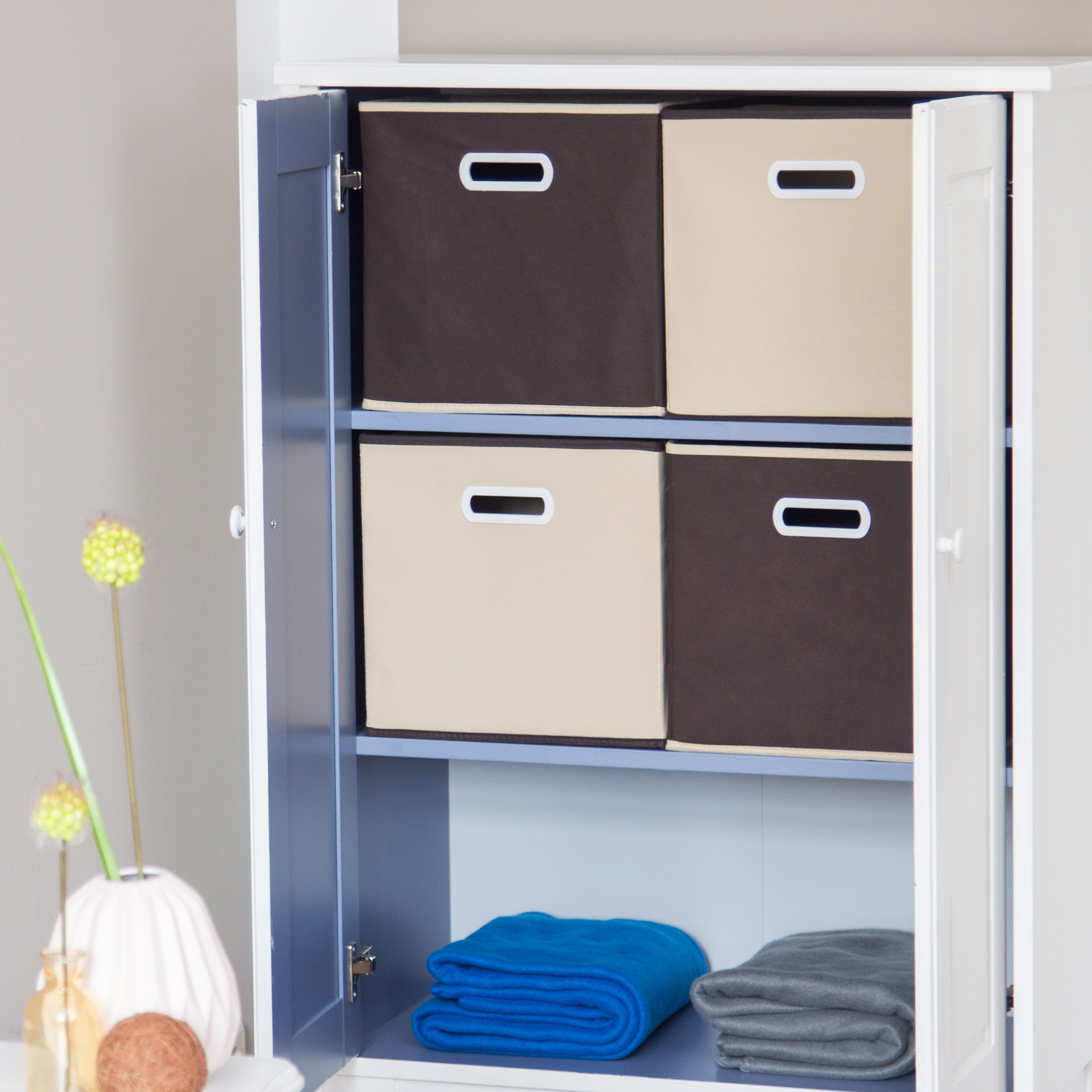 Weißer Schrank mit offenen Türen, braunen und beige Kisten oben und gefalteten blauen und grauen Decken unten.