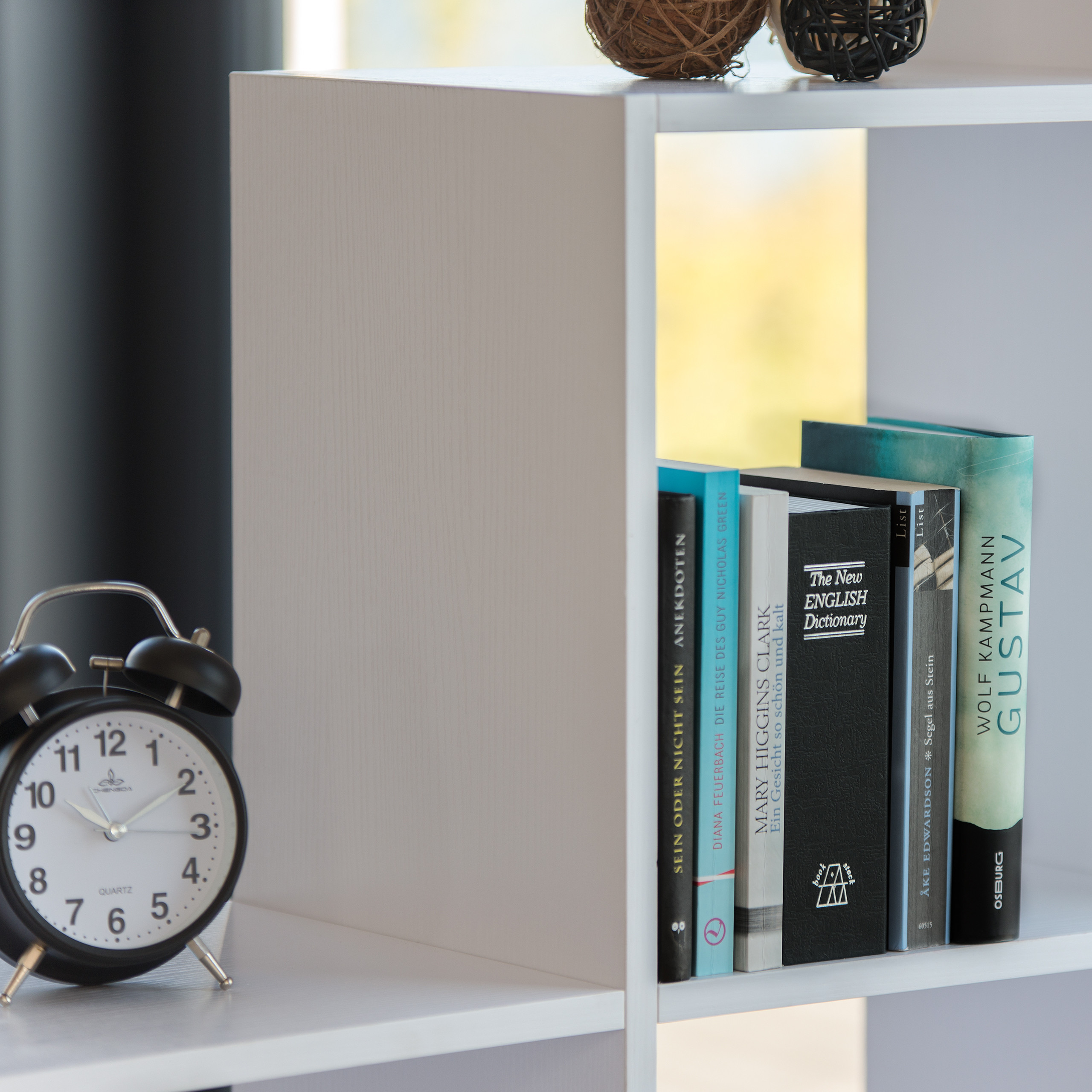 Ein schwarzer Wecker steht auf einem weißen Regal neben einem Fach mit vertikalen Büchern.