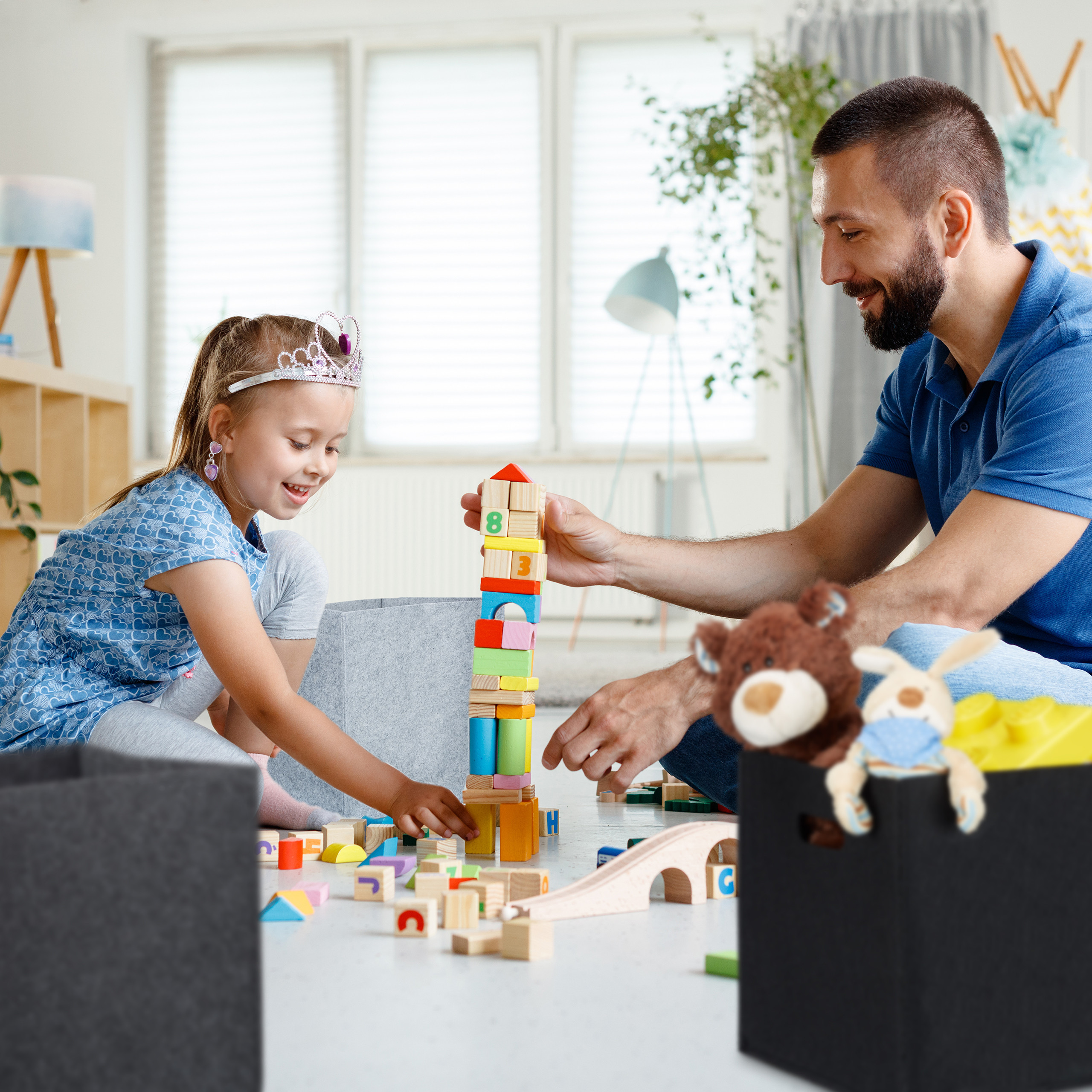 Ein Vater und seine kleine Tochter im Diadem bauen zusammen im hellen Wohnzimmer einen bunten Holzklotzturm.
