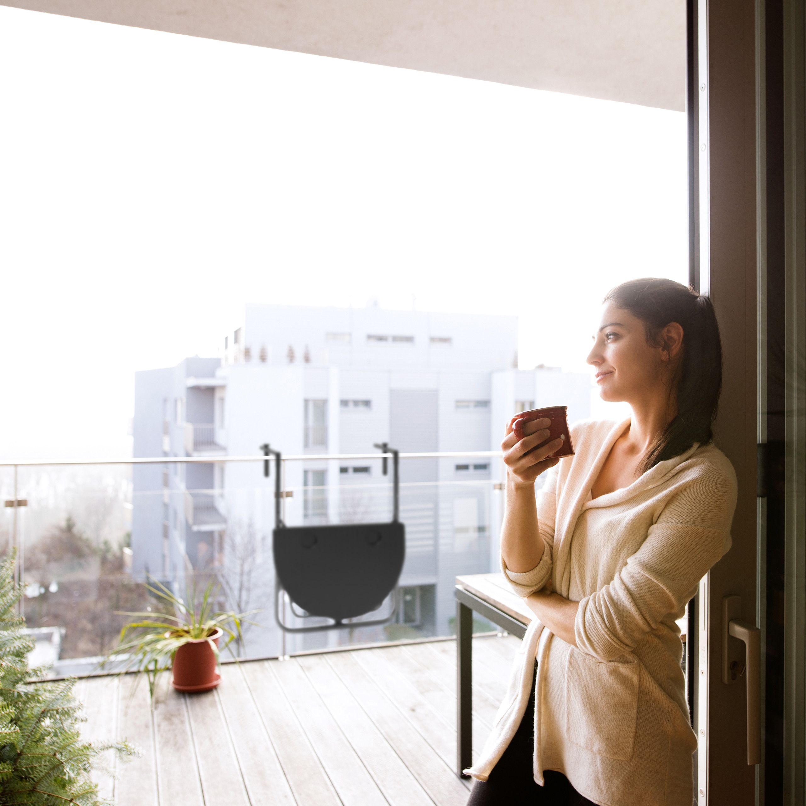 Frau mit einer Tasse Kaffee steht auf einem sonnigen Balkon mit Stadtblick.
