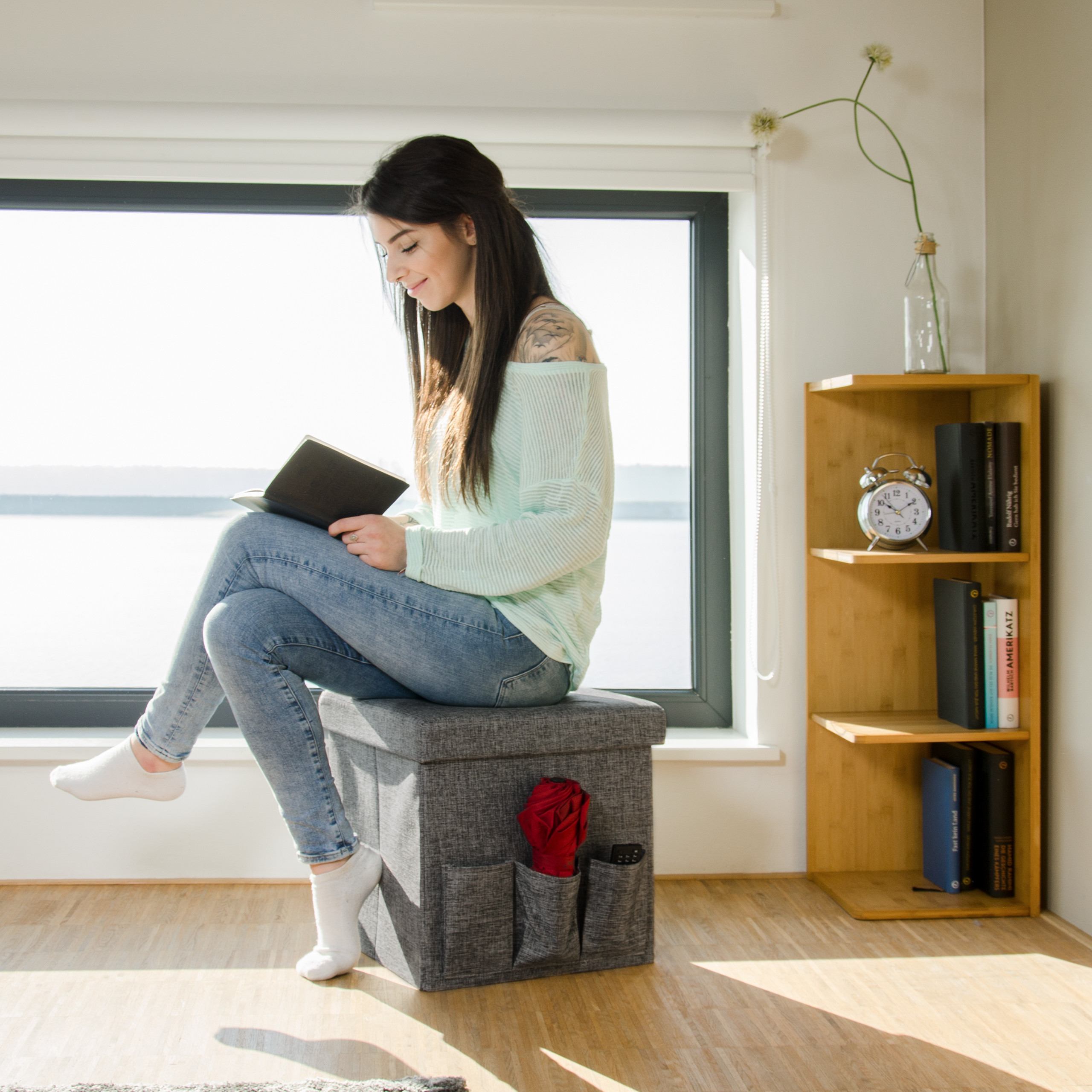 Eine Frau sitzt auf einem grauen Hocker und liest ein Buch vor einem grossen Fenster.