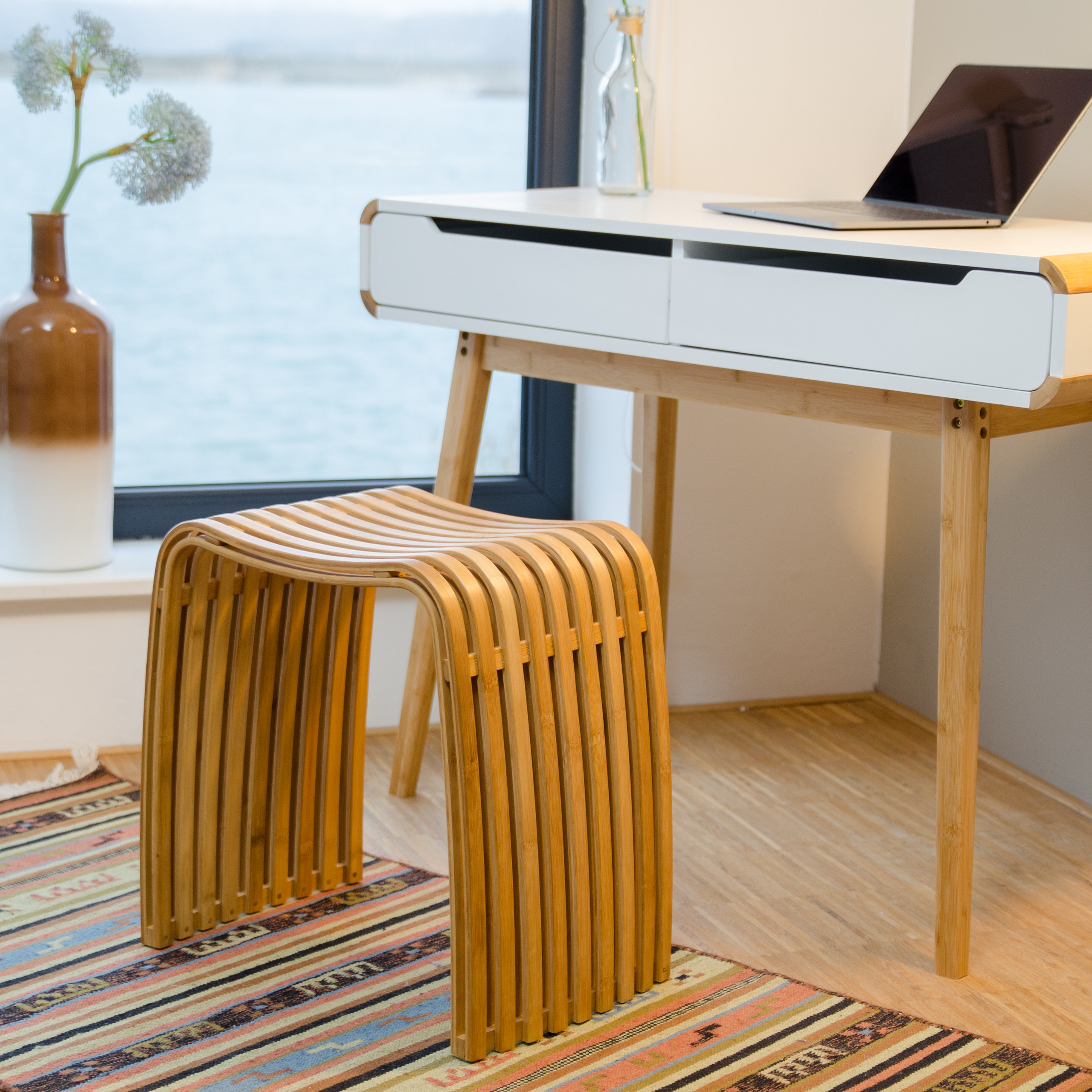 Ein weißer Holztisch mit Laptop steht an einem Fenster, davor ein geschwungener Hocker auf einem Teppich.