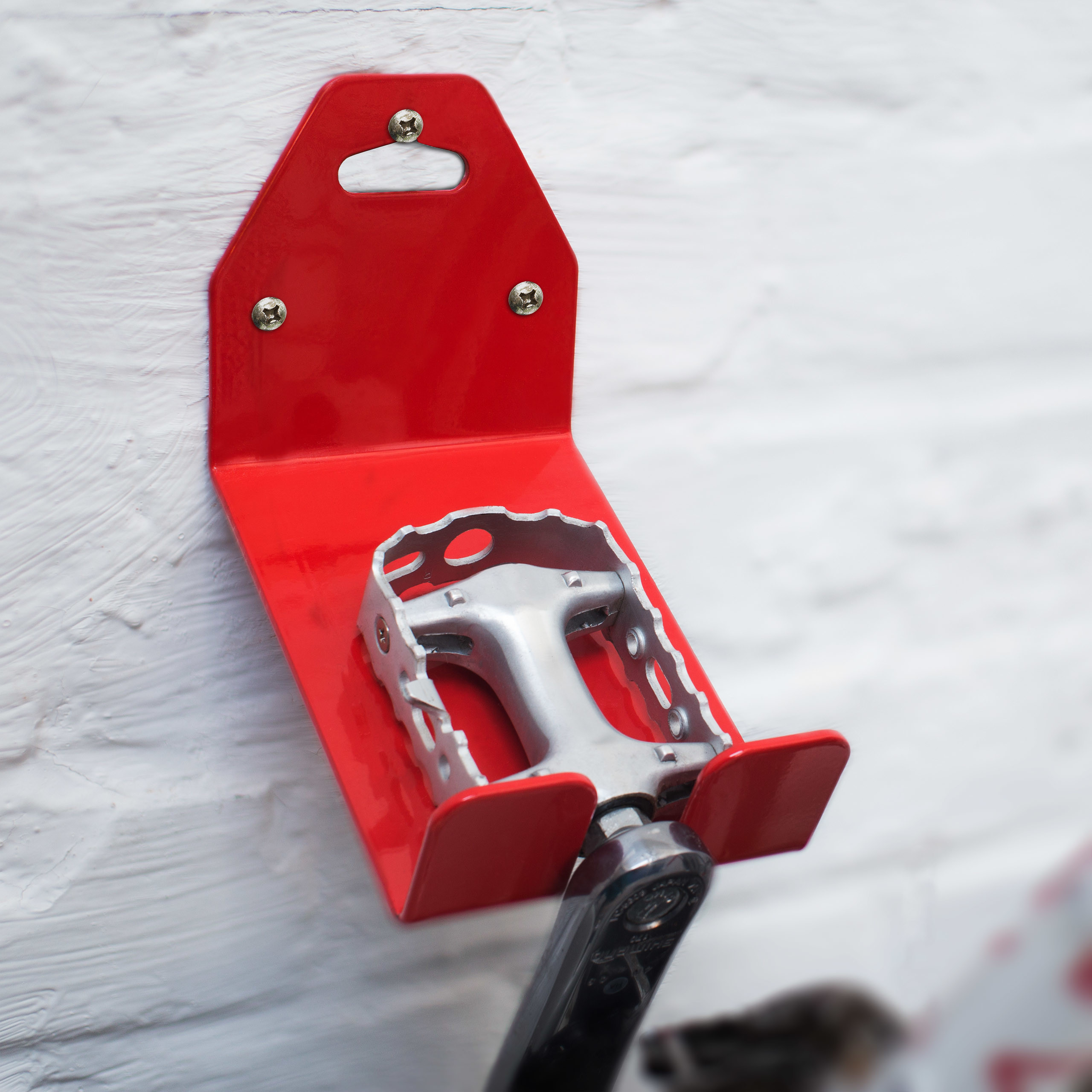Eine rote Wandhalterung aus Metall hält ein silbernes Fahrradpedal an einer weißen Backsteinwand.