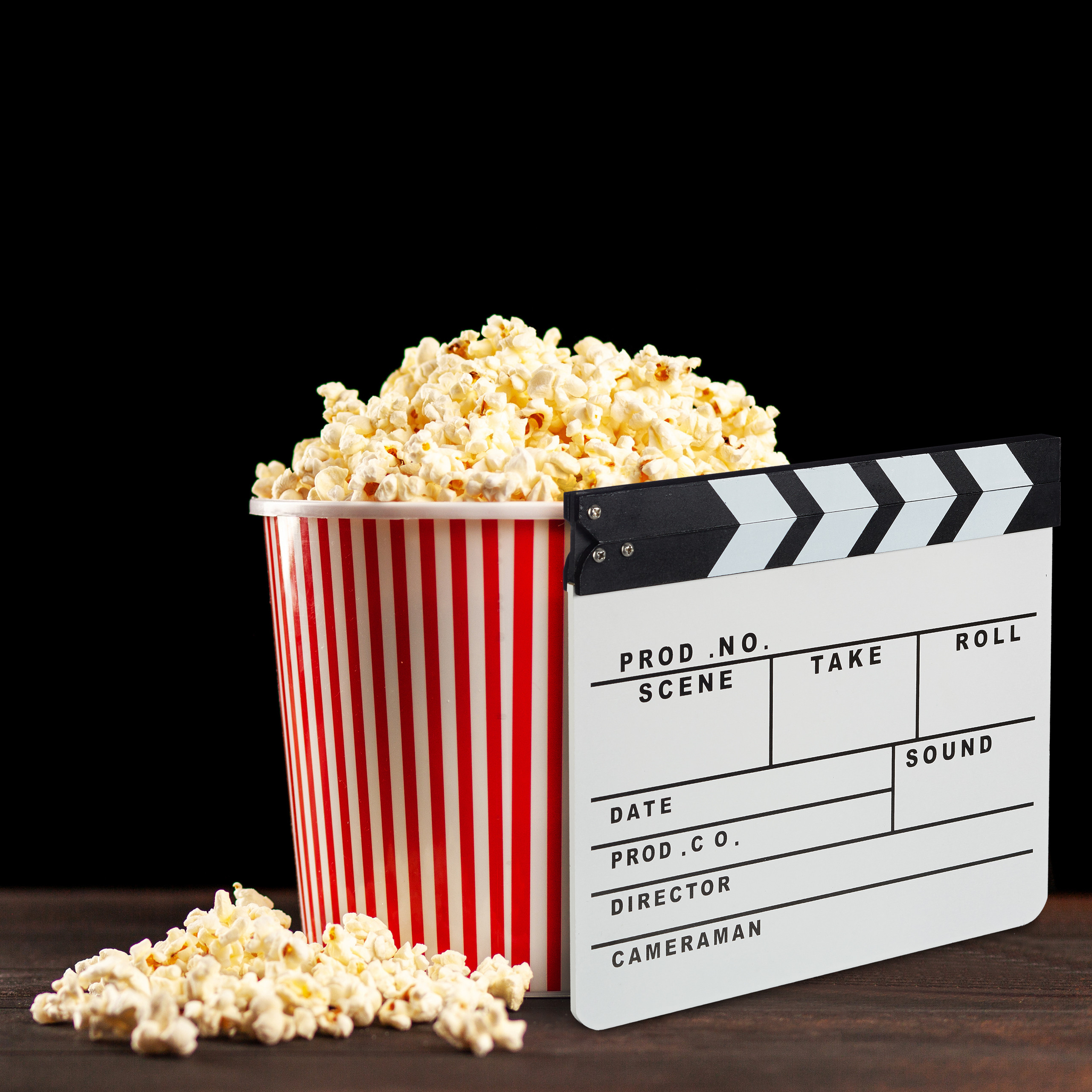 Eine rot-weiß gestreifte Popcorntüte mit Popcorn und eine Filmklappe stehen auf Holz vor schwarzem Hintergrund.